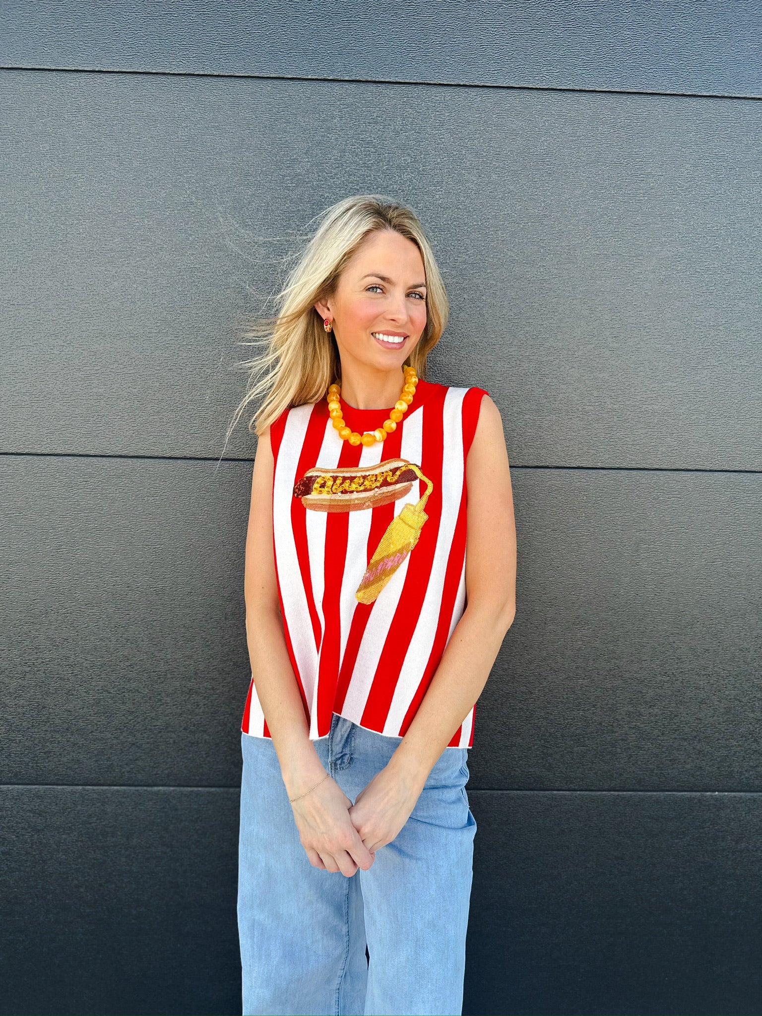 Woman wearing a red and white striped knit tank top with a sequined hot dog graphic, paired with light wash jeans.