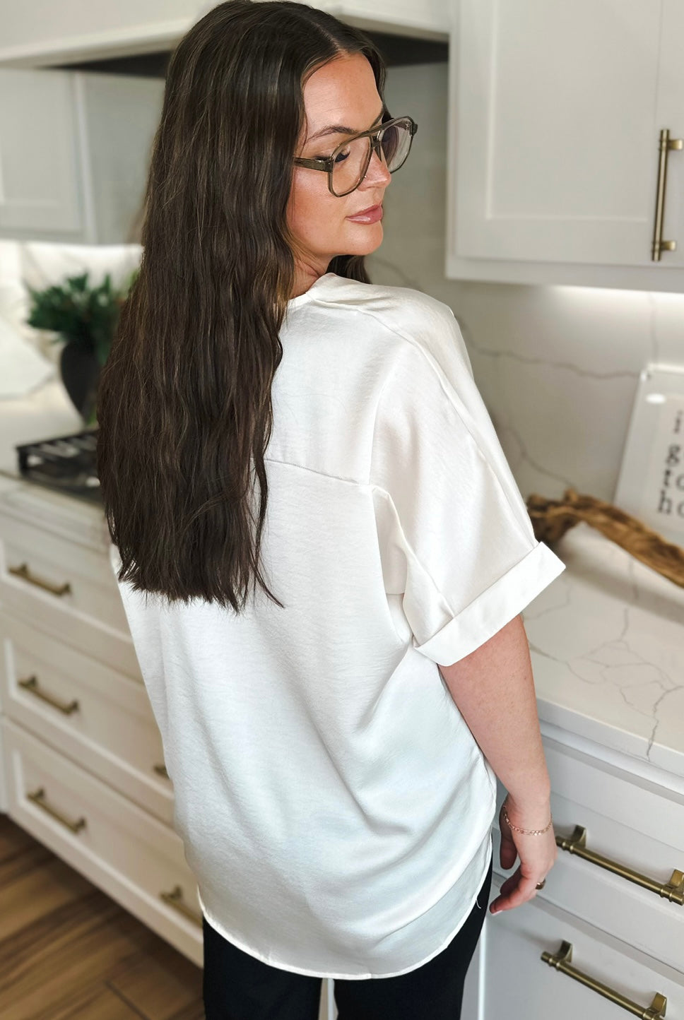 Woman from behind wearing an ivory satin v-neck high-low blouse with cuffed short sleeves. She has long brown hair and