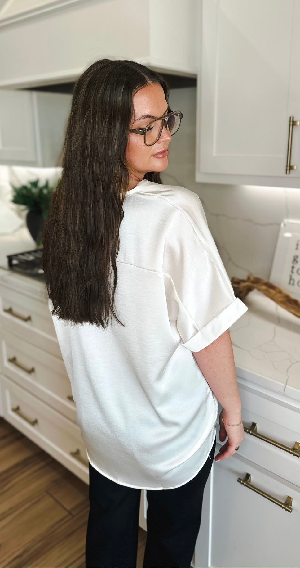 Woman from behind wearing an ivory satin v-neck high-low blouse with cuffed short sleeves. She has long brown hair and
