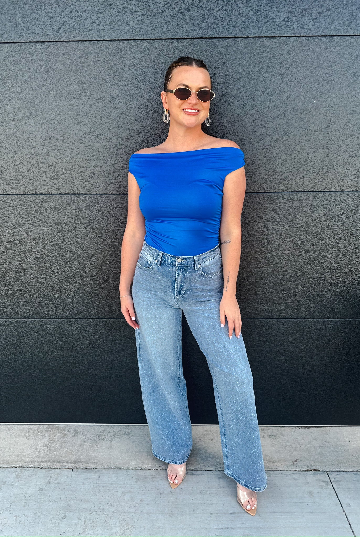 Woman wearing light wash, slouch wide-leg jeans and a bright blue off-the-shoulder top, sunglasses, and hoop earrings.
