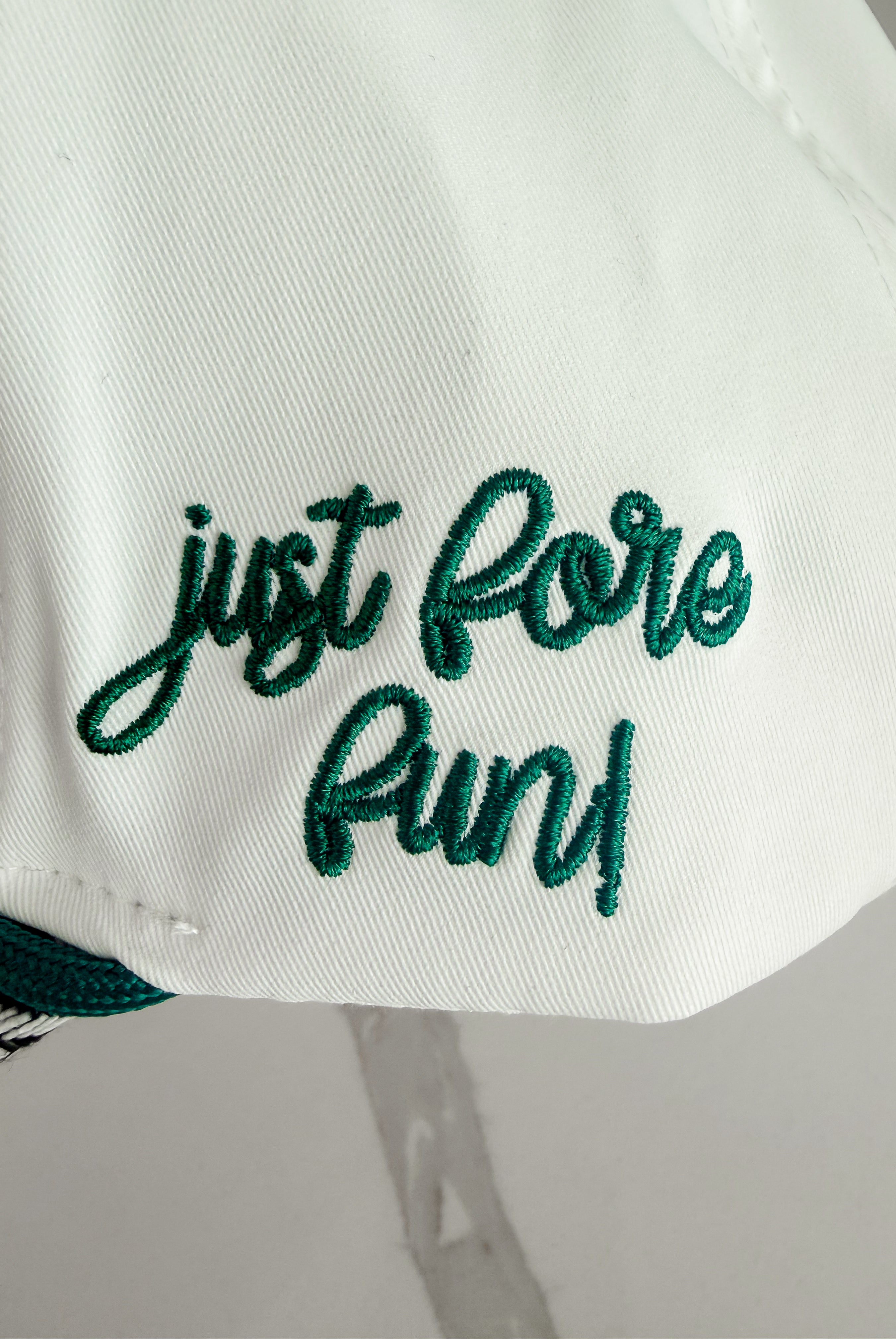 White caddie hat with "just fore fun!" embroidered in green script. Herringbone pattern visible on brim.