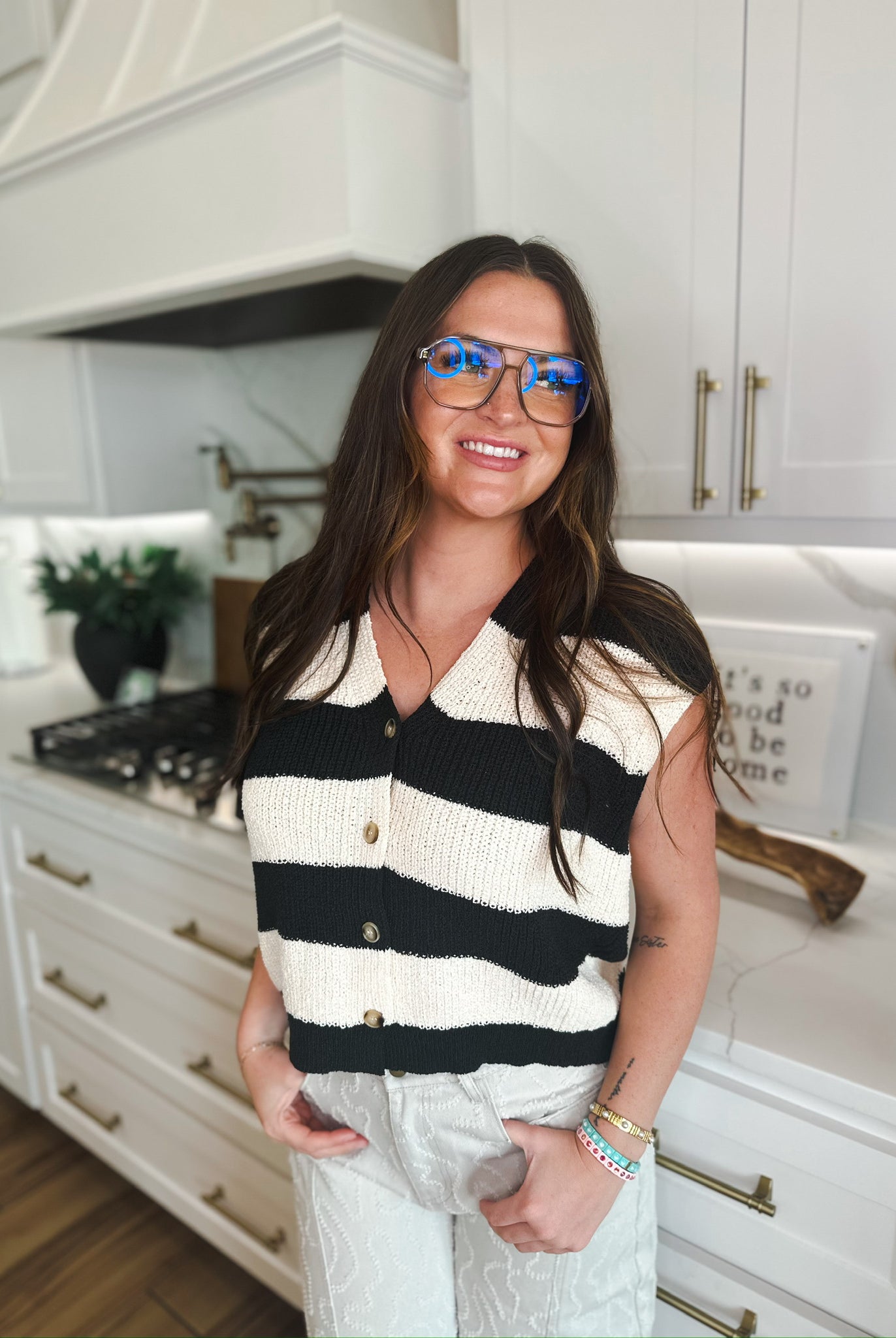 Woman wearing a black and cream striped knit sweater vest with v-neck and button detail, paired with textured silver pants.