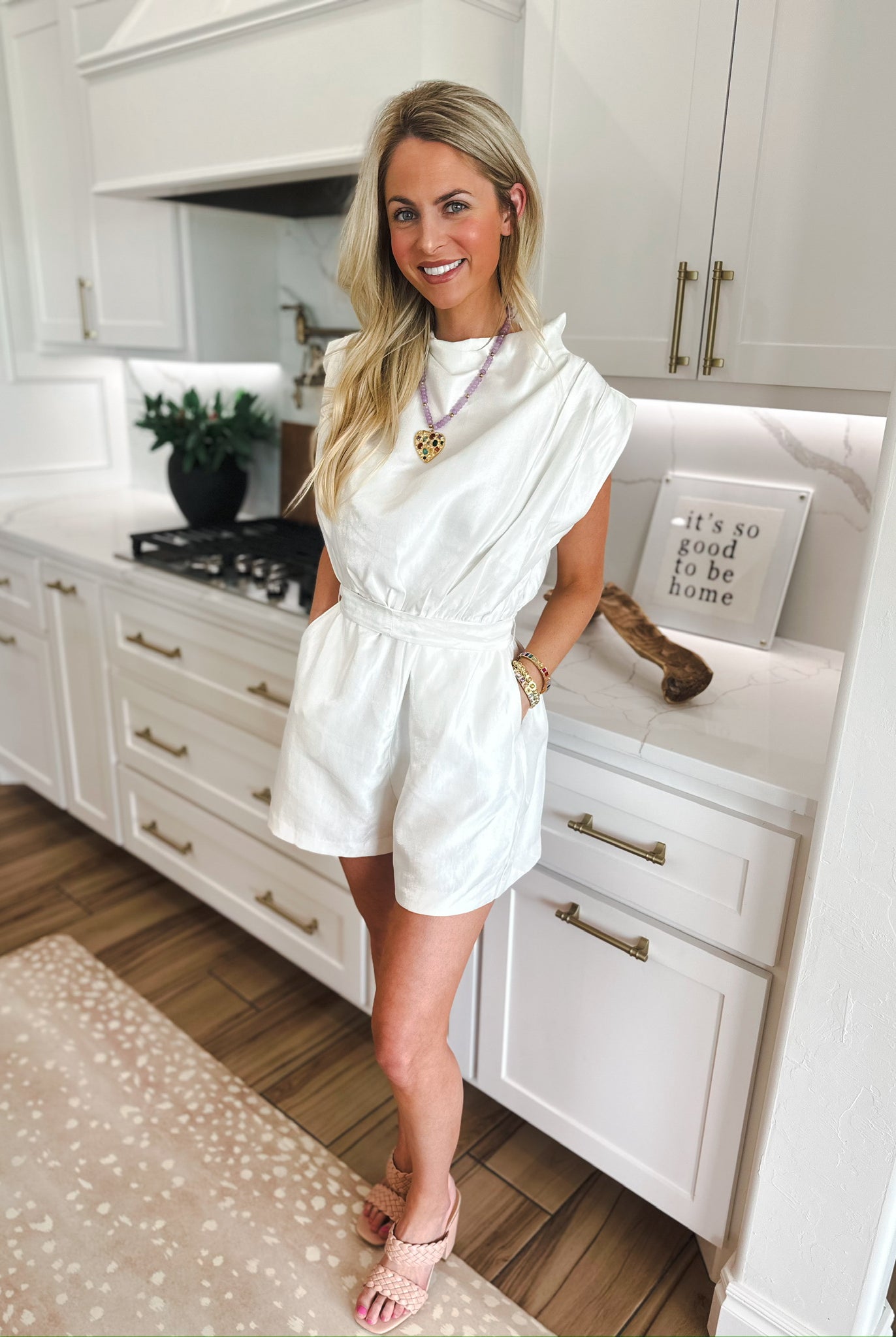 Woman in white shimmer romper with cowl neck and cap sleeves. Romper has a belted waist and pleated shorts.