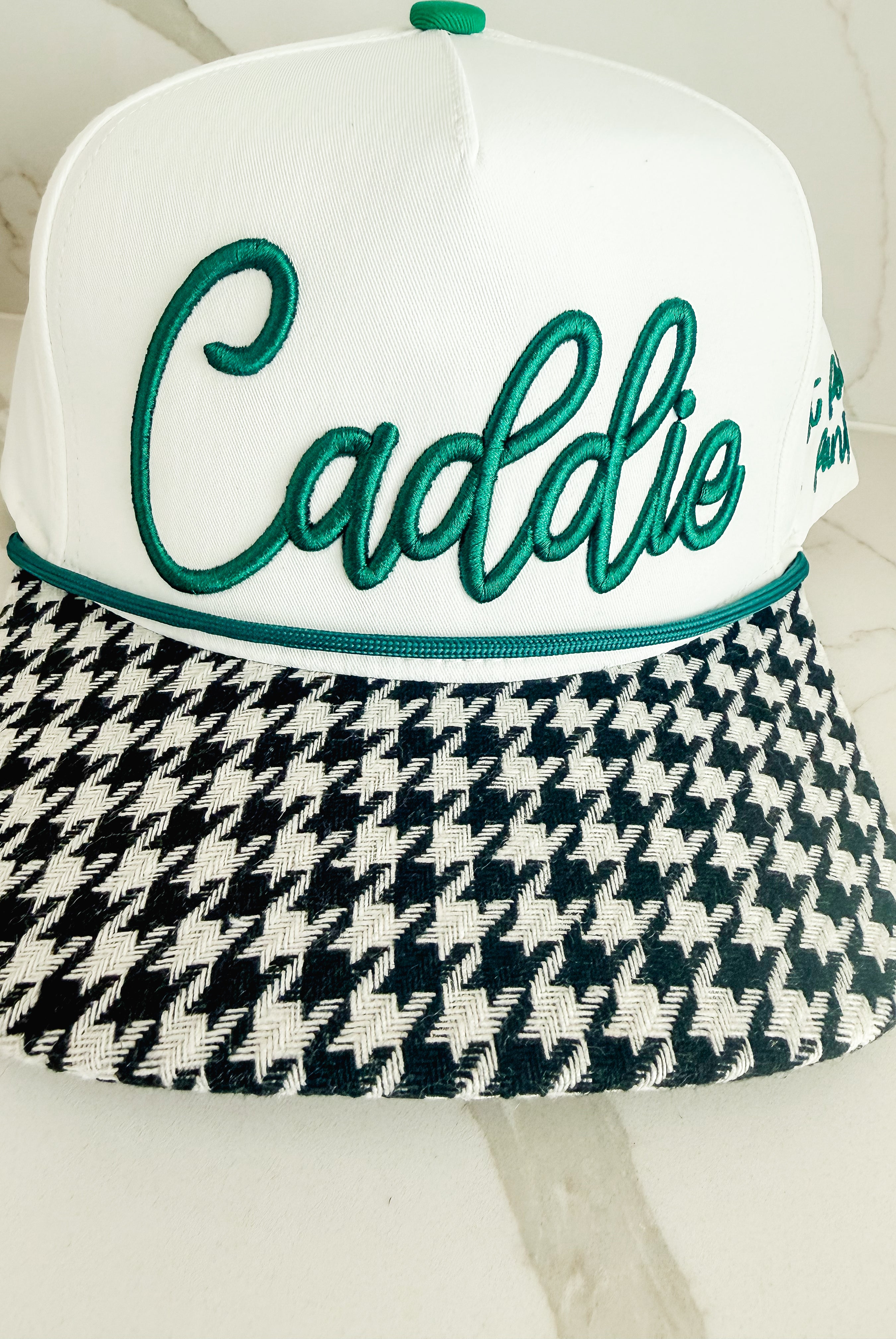 White caddie hat with green embroidered script logo and black and white houndstooth brim. Green rope accent above brim.