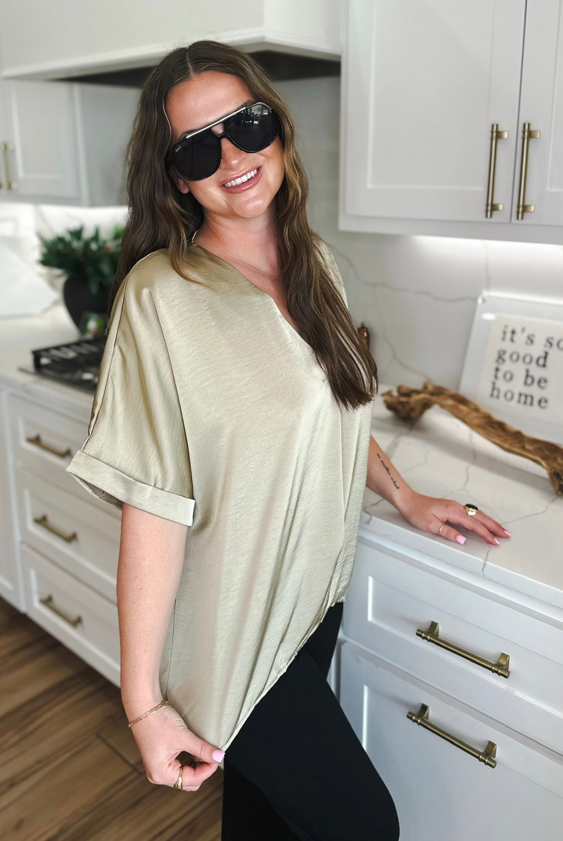 Woman wearing a taupe satin V-neck blouse with cuffed short sleeves and black leggings. She has on large black sunglasses