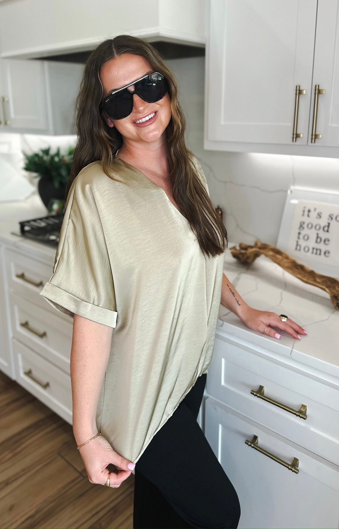 Woman wearing a taupe satin V-neck blouse with cuffed short sleeves and black leggings. She has on large black sunglasses