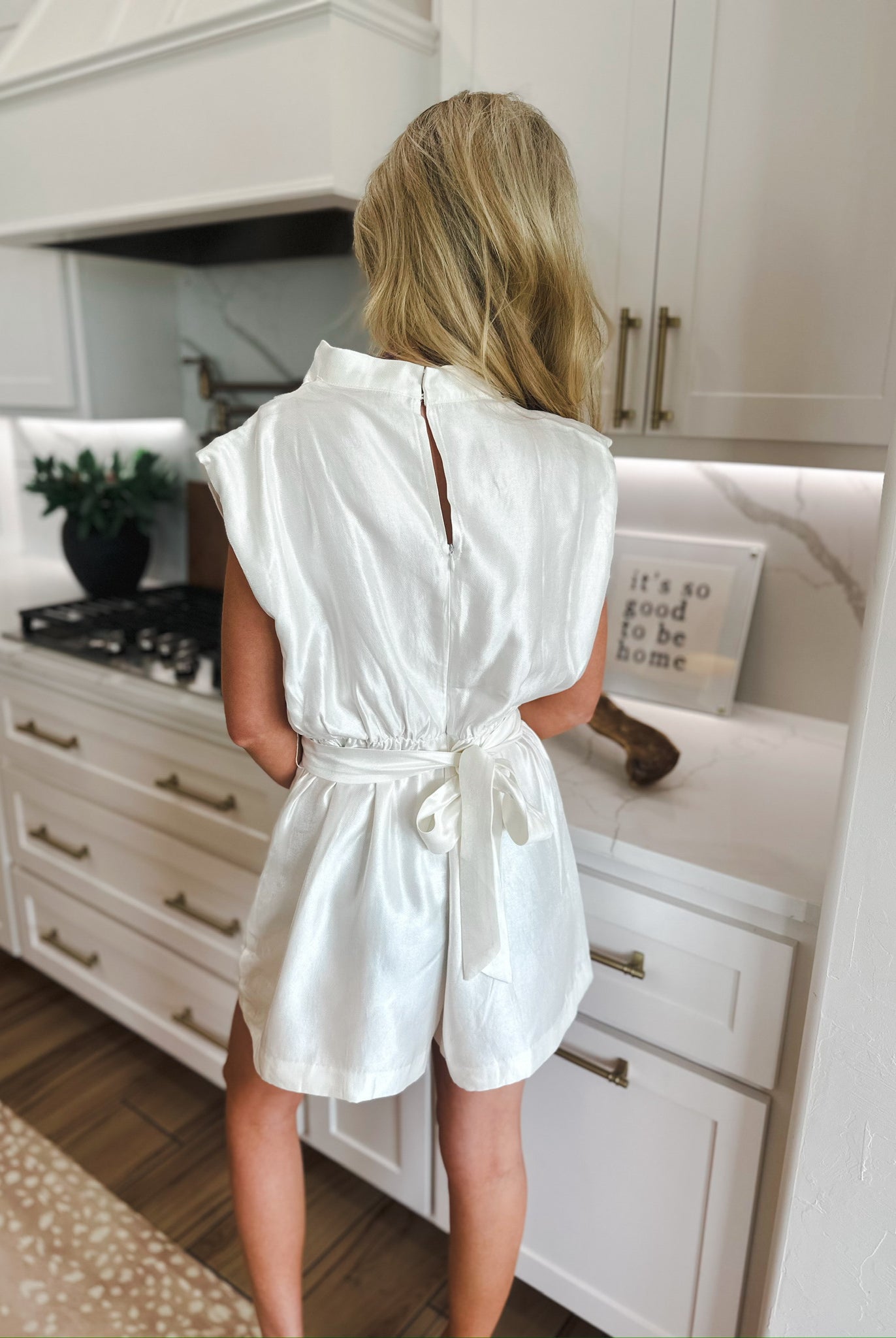 Back view of a woman wearing a white shimmer cowl neck romper with a sash tie at the waist and a keyhole back closure.