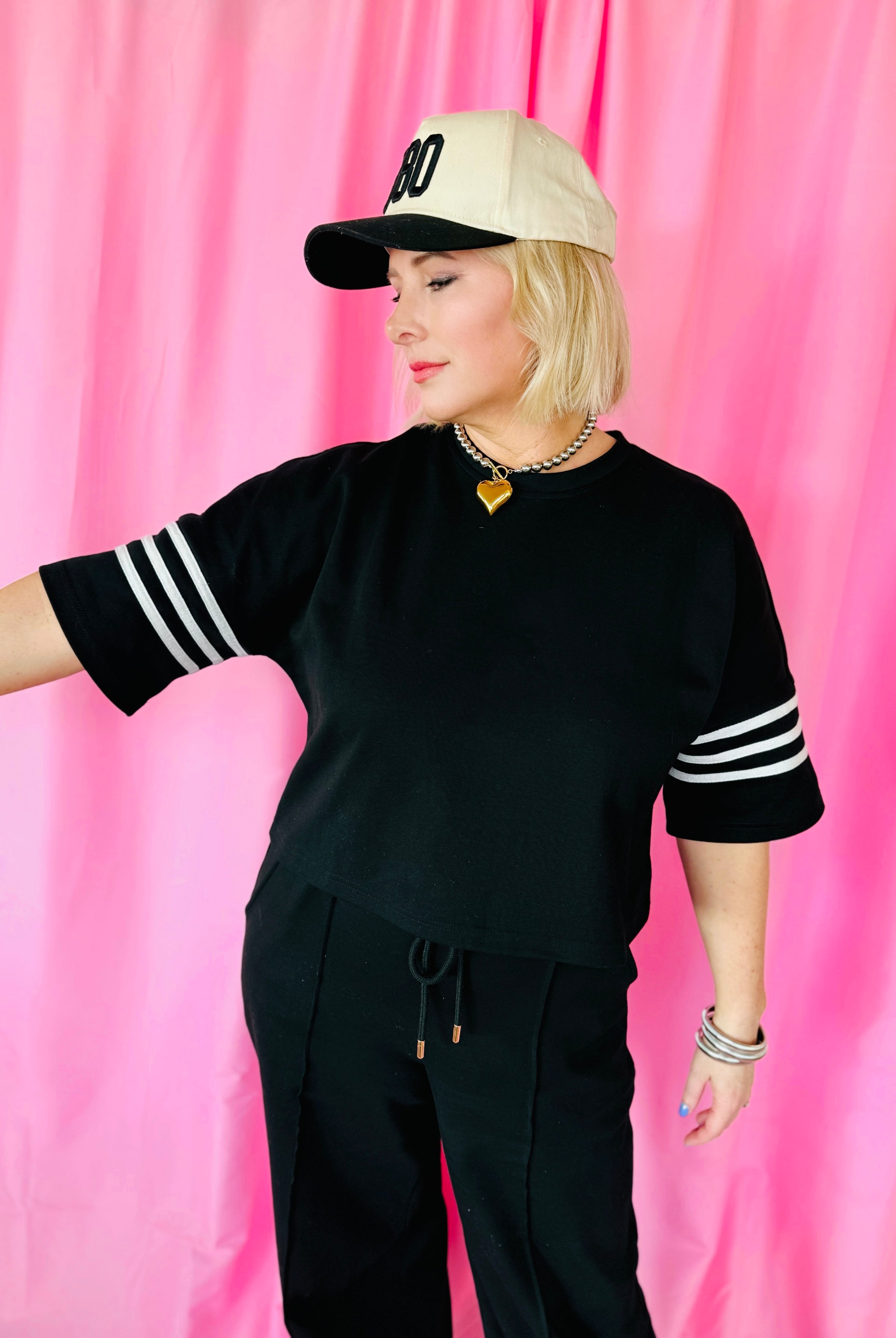 Woman wearing a black short-sleeve top with white stripes on the sleeves, black drawstring pants, and a beige baseball cap