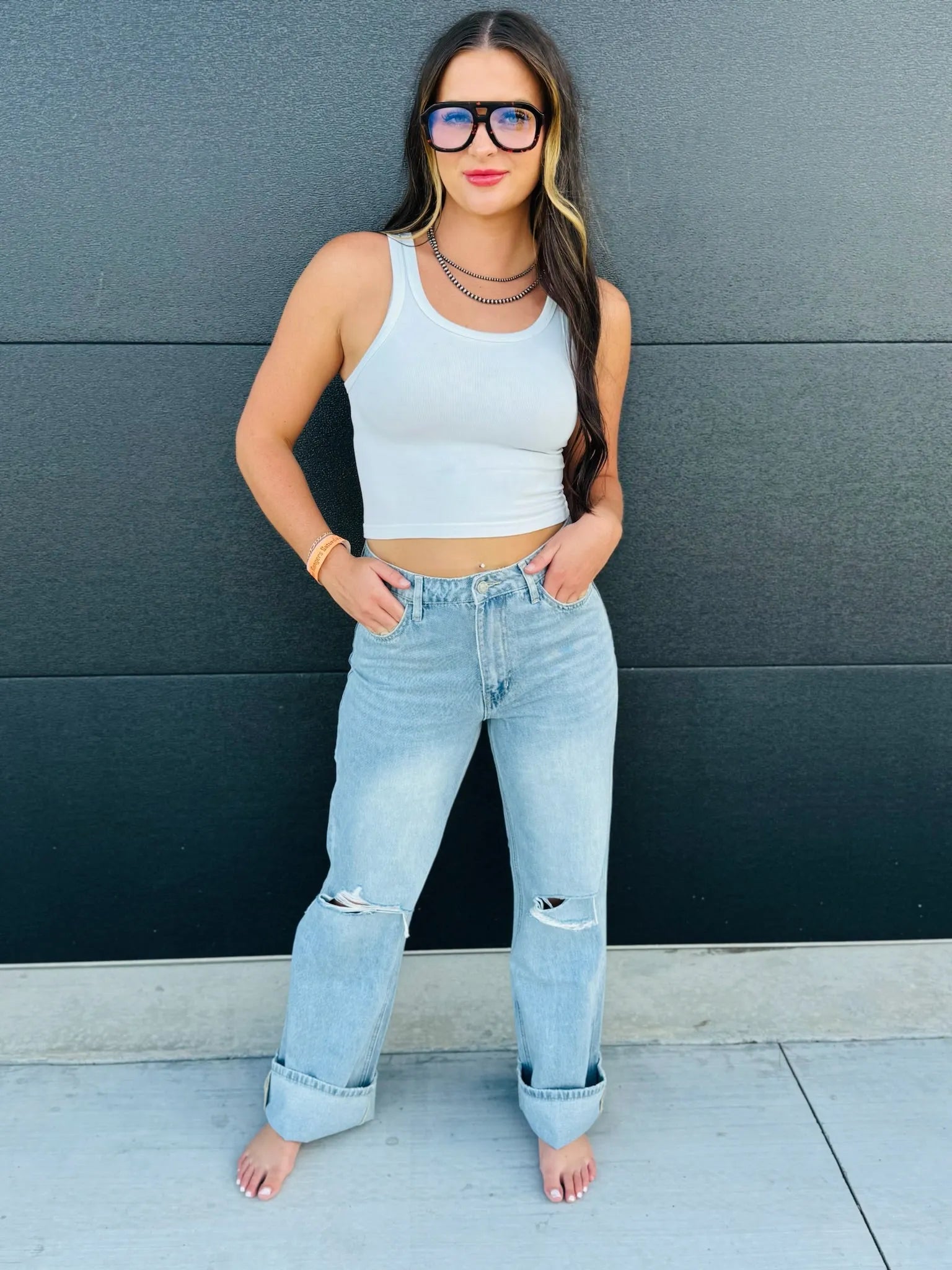 Woman in light blue high-rise flare jeans with ripped knees and cuffed hems, wearing a white tank top and large