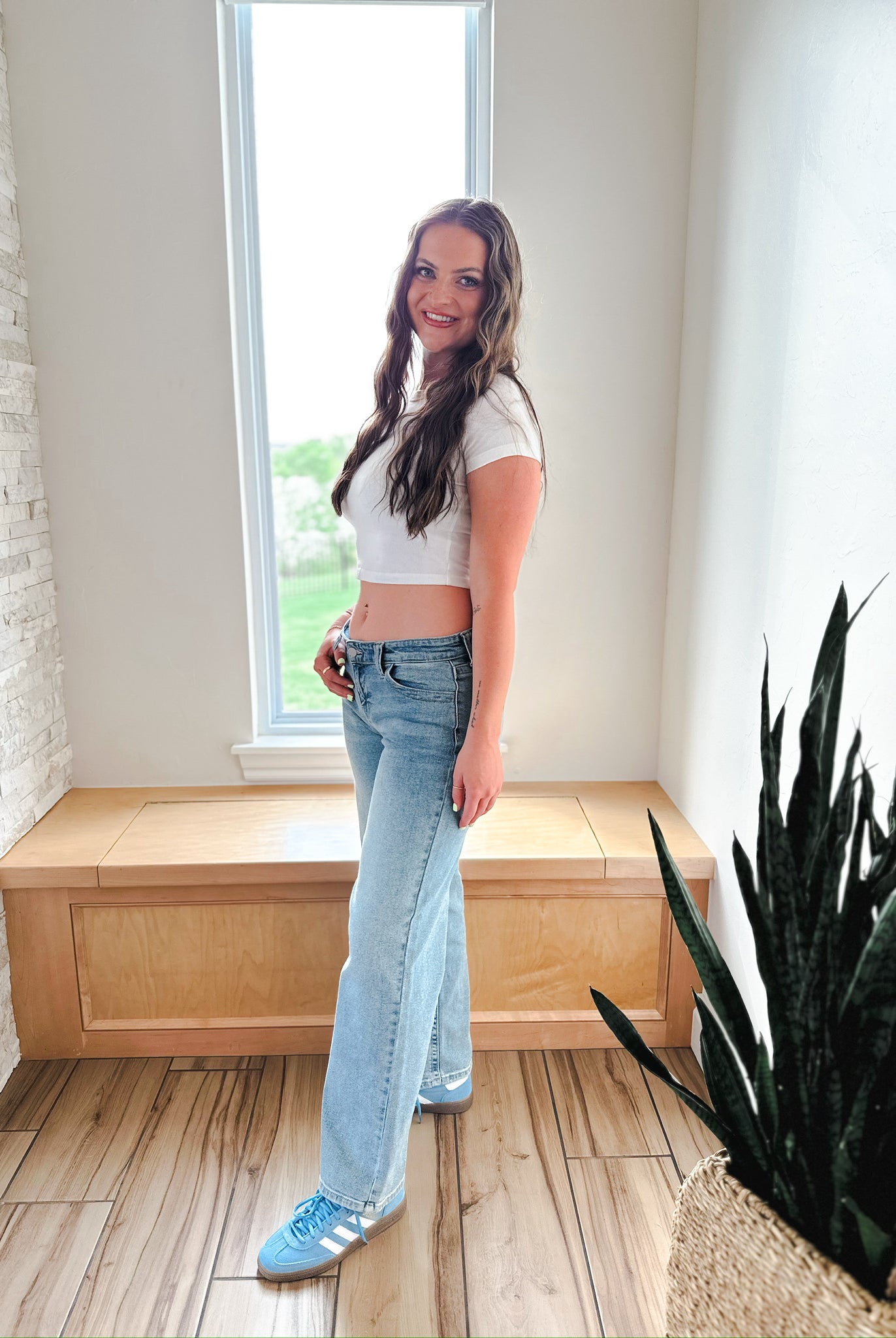 Model wearing JBD wide leg medium wash low rise denim jeans and a white crop top, standing indoors near a window.