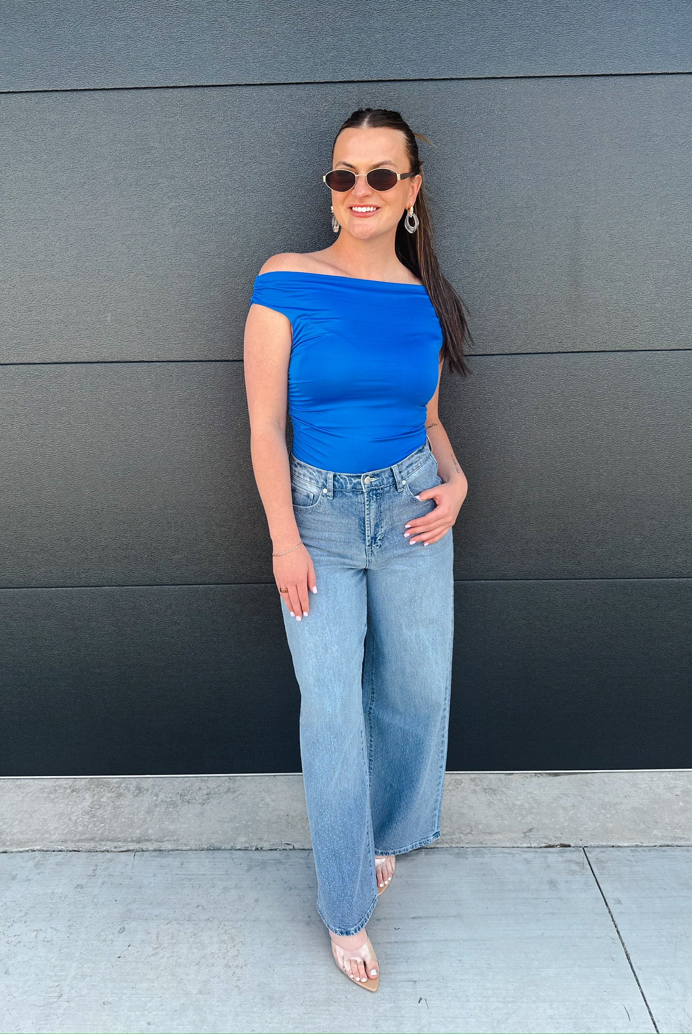 Woman wearing Habitual Gemma wide-leg jeans, a blue off-the-shoulder top, sunglasses and earrings, standing against a dark