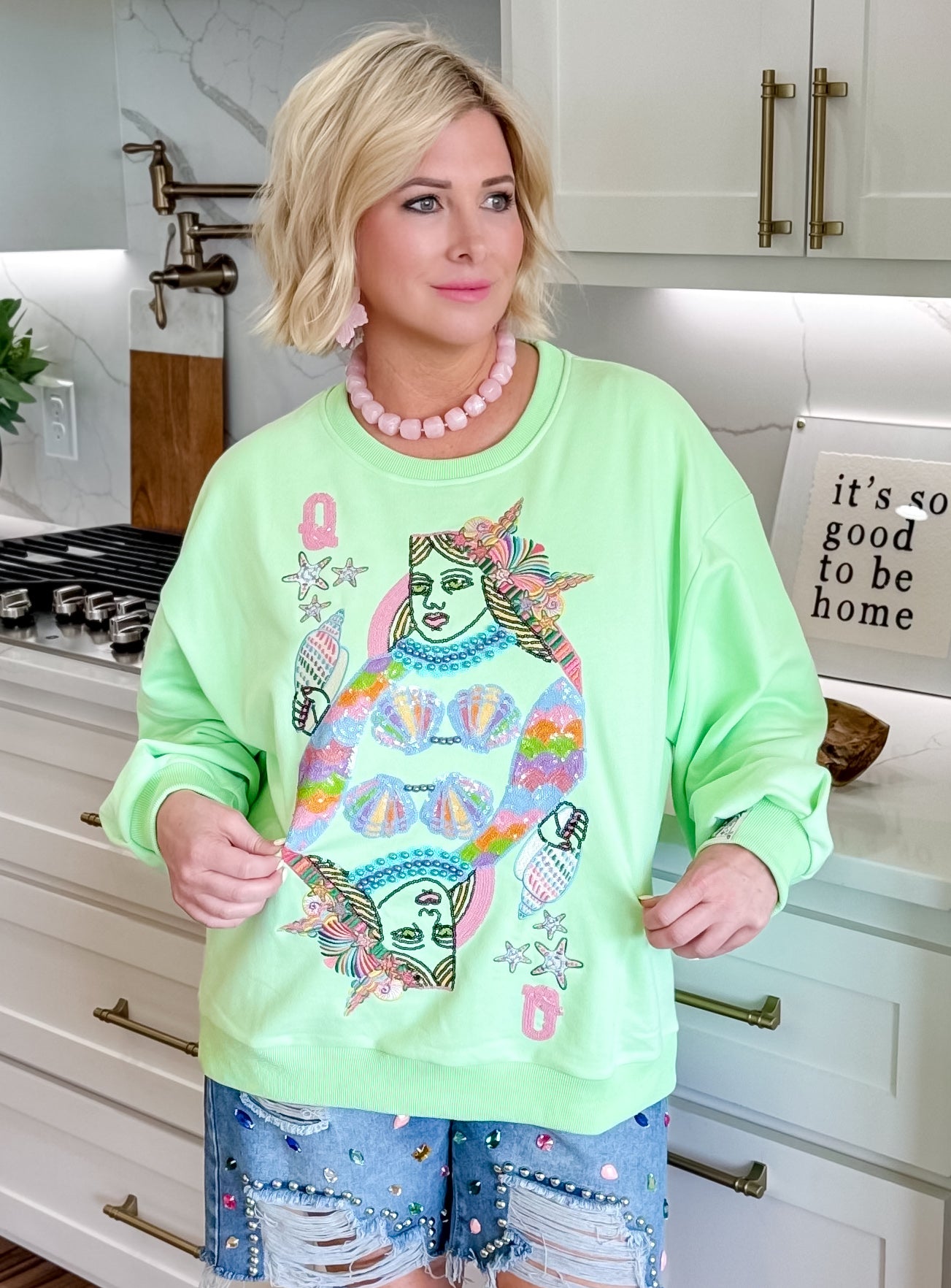 Light green sweatshirt with a "Queen of the Sea" card design featuring a sequined mermaid, shells, and stars. Woman wearing