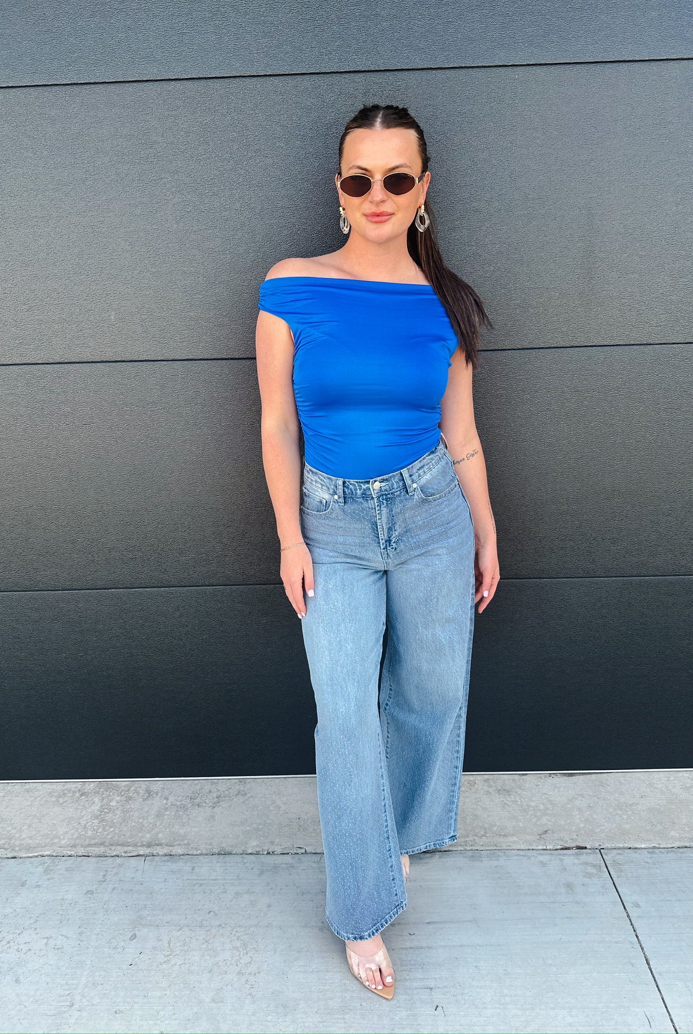 Model wears light wash slouch wide leg jeans with an off-the-shoulder royal blue top, sunglasses, and clear heels against a