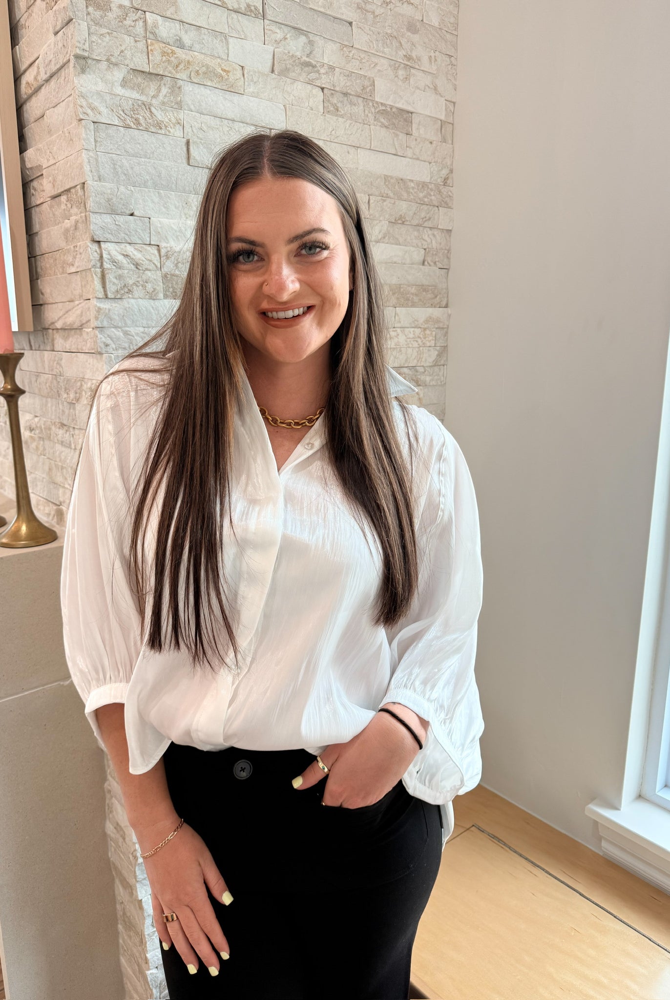 Woman wearing a white shiny button-front blouse with wide, long sleeves and a gold chain necklace, paired with black pants.
