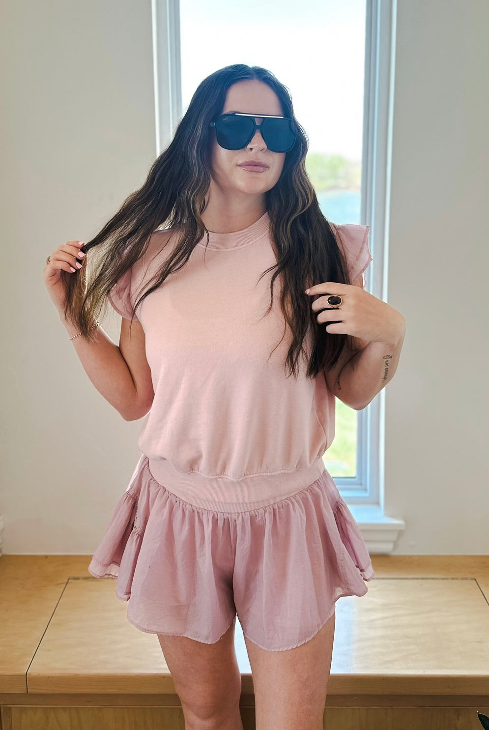 Woman in light pink crinkle nylon athleisure set featuring ruffle sleeve pullover and flowy shorts. She wears large black