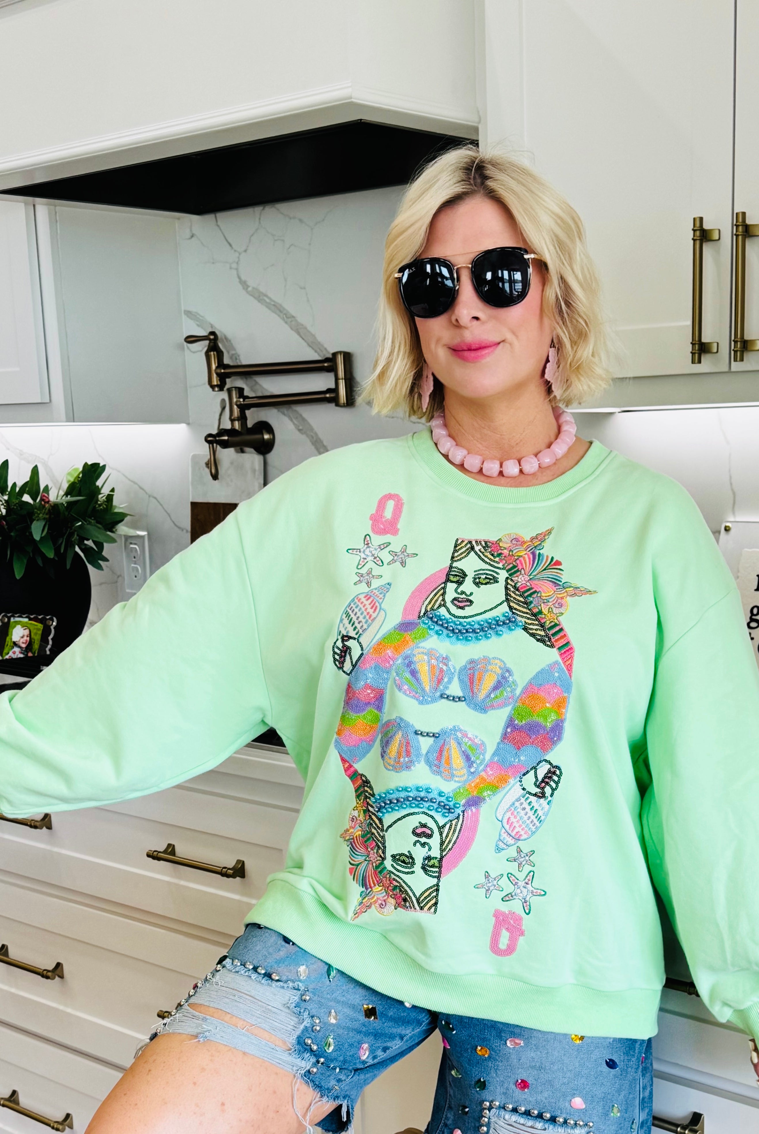 Light green Queen of the Sea sweatshirt with a graphic of two queens' faces and colorful seashell details. Woman wears