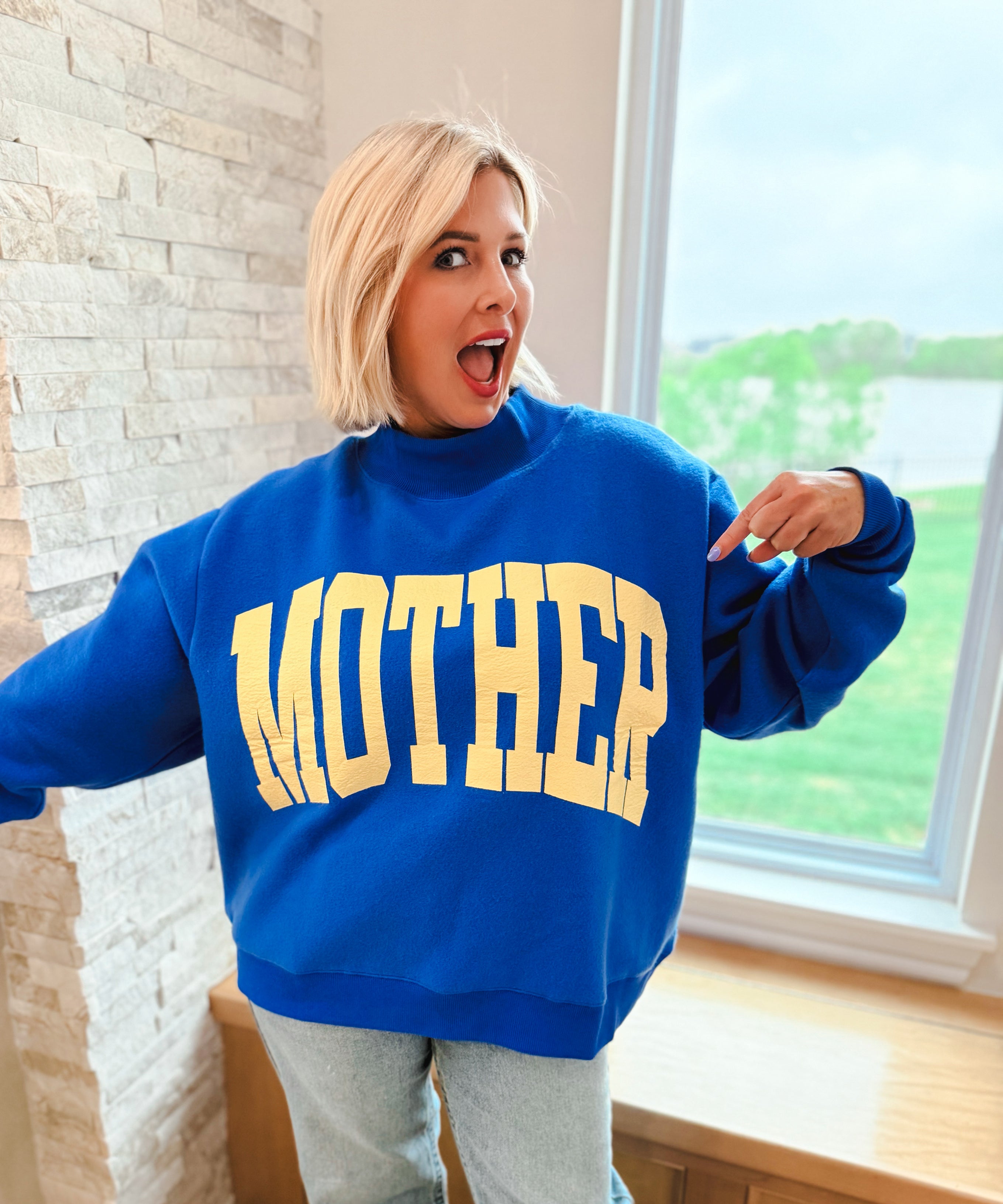 Oversized royal blue mockneck sweatshirt with "MOTHER" in gold varsity letters, worn by a woman.