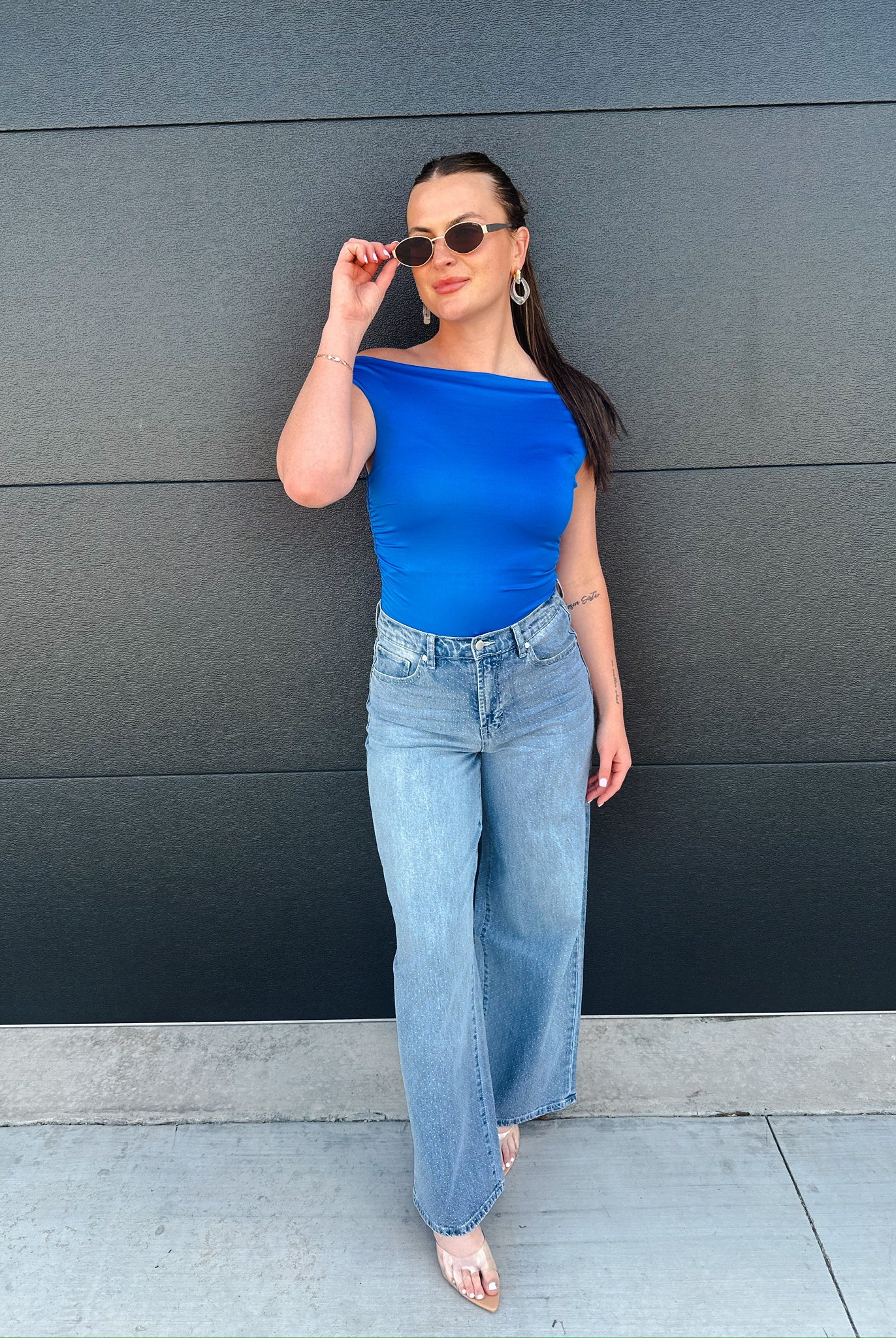 Woman in blue off-the-shoulder top and light wash wide-leg jeans, posing against a dark gray wall.