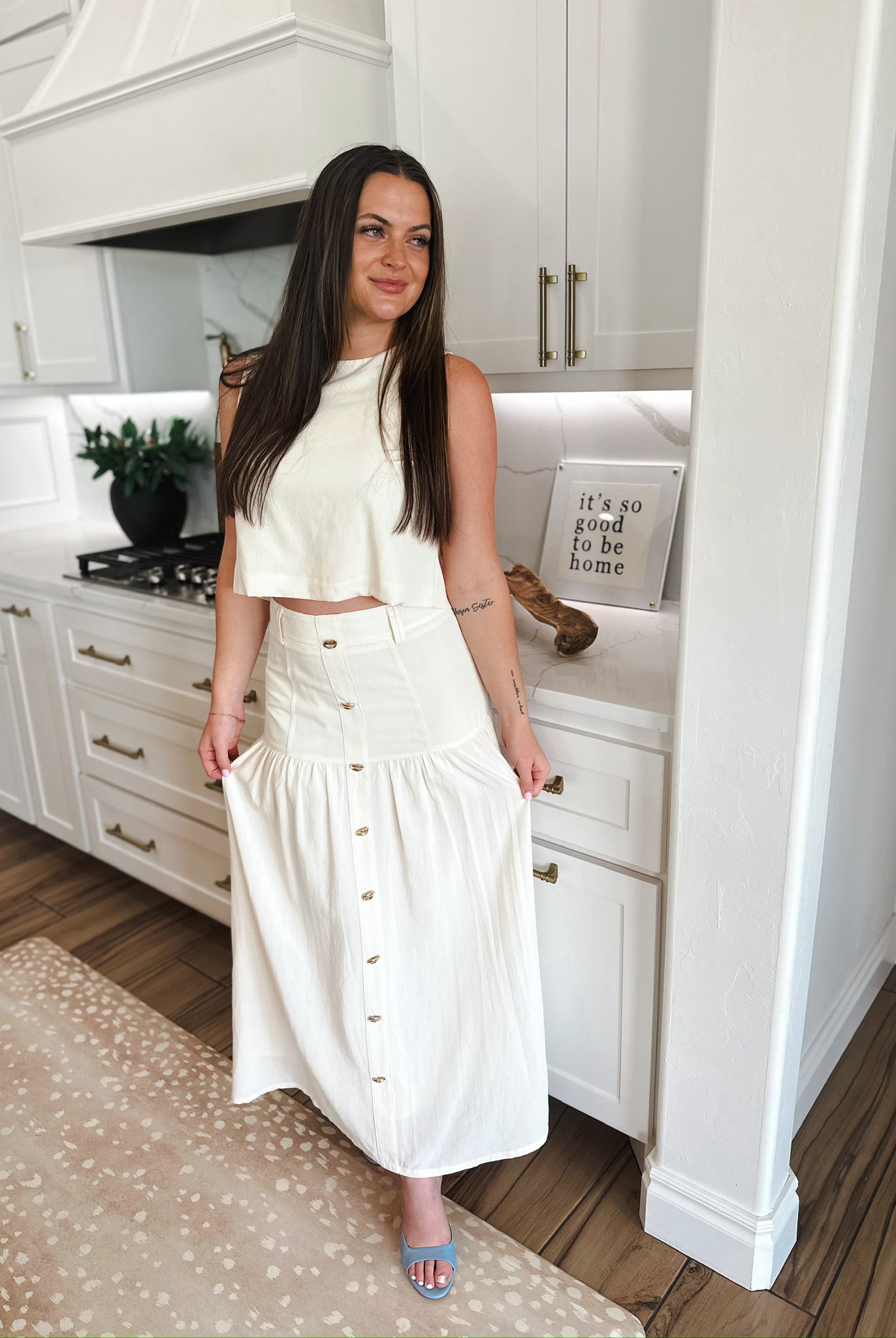 Woman in elegant cream matching set: cropped top and button-front maxi skirt. Kitchen setting.