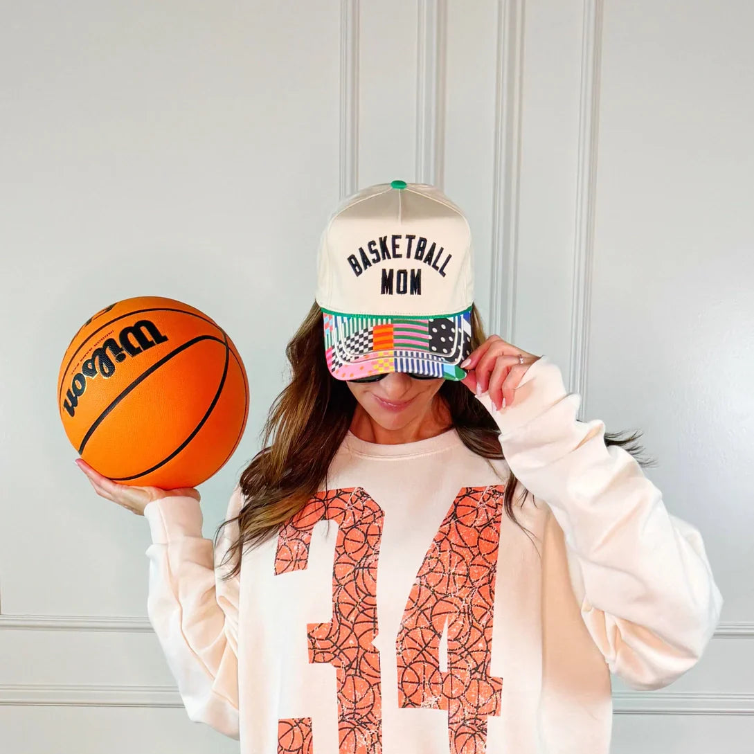 Woman in a cream sweatshirt with '34' basketball print, wearing a beige trucker hat with "BASKETBALL MOM" and a colorful