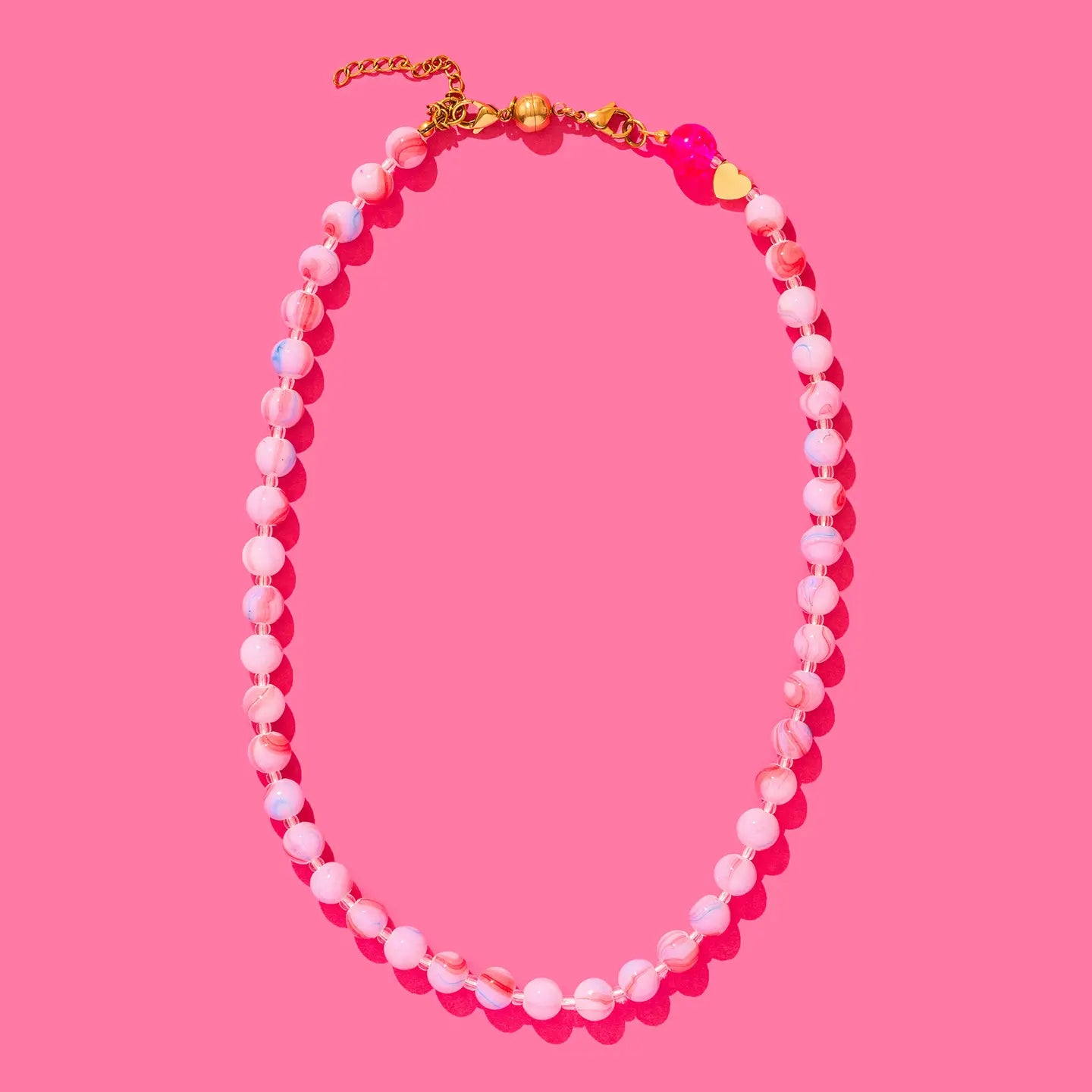 Beaded layering necklaces with pink and white marbled beads, a gold chain, and a pink heart charm on a pink background.