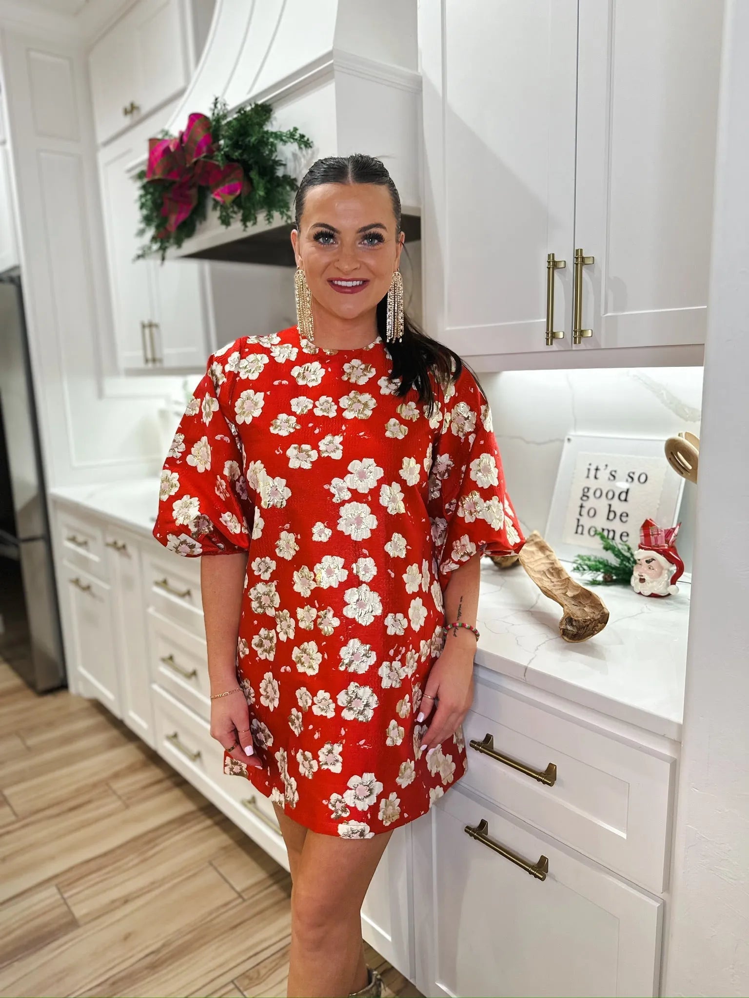 Woman in red jacquard short dress with white floral print, puff sleeves, and statement earrings in a modern kitchen.