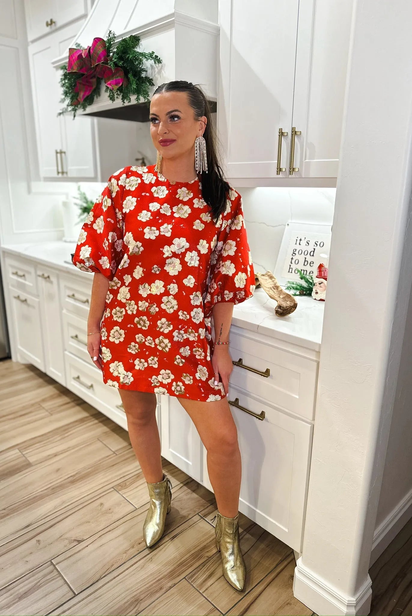 Woman in a red floral jacquard short dress, gold metallic ankle boots, and long crystal earrings, standing in a kitchen.