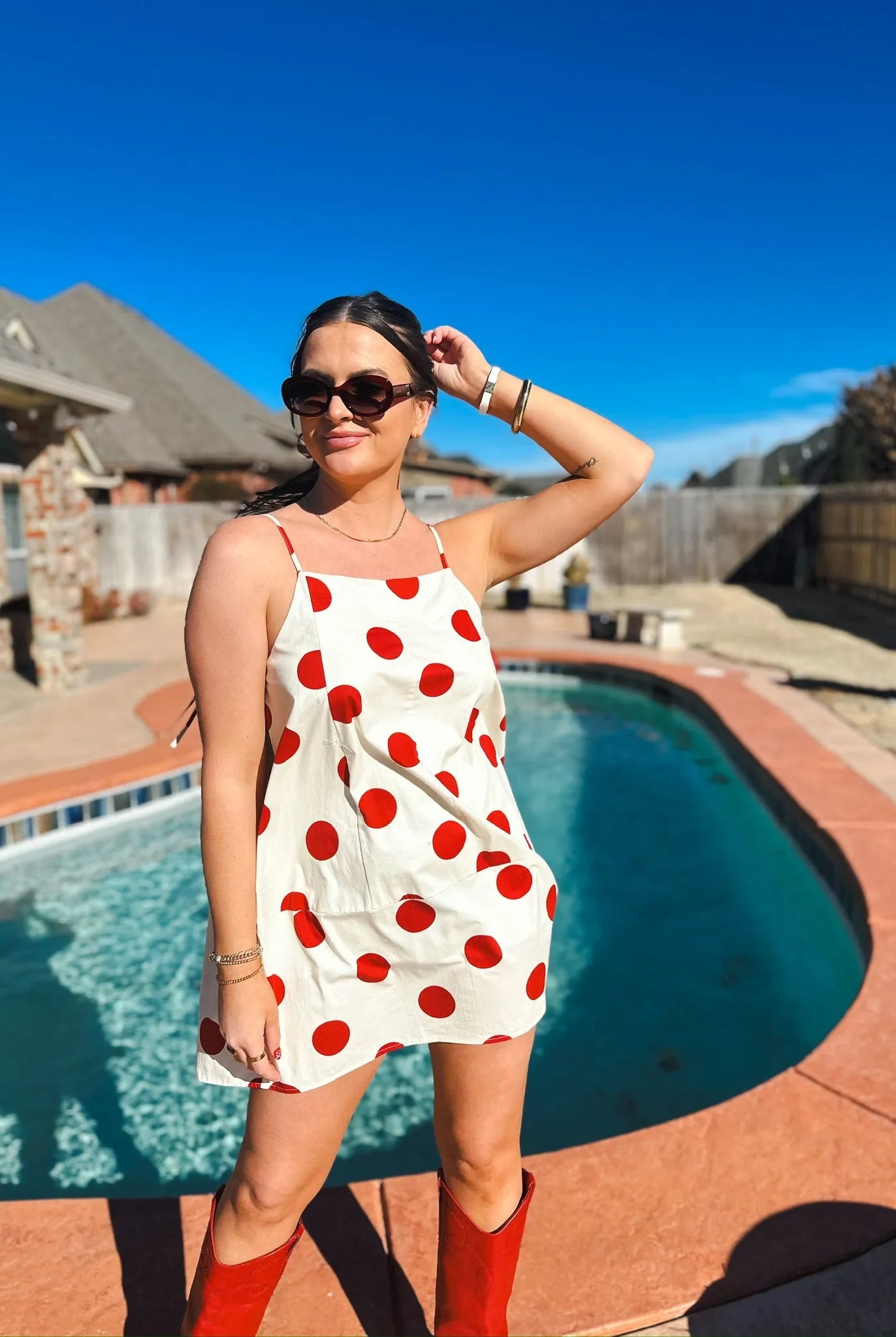 Woman in red and cream polka dot mini dress with spaghetti straps, red boots, sunglasses. Poolside setting.