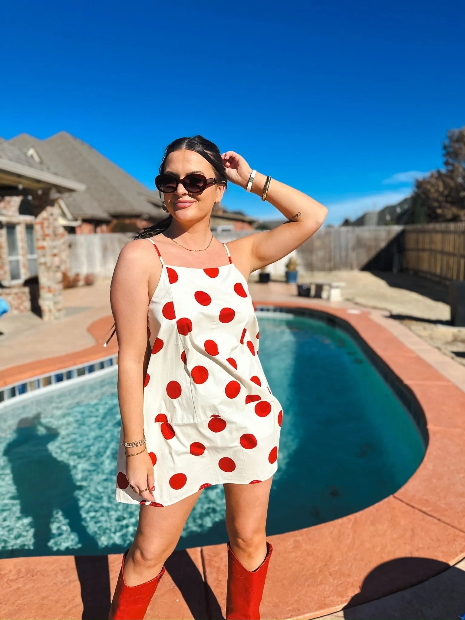 Woman in red and cream polka dot mini dress with spaghetti straps, red boots, sunglasses. Poolside setting.