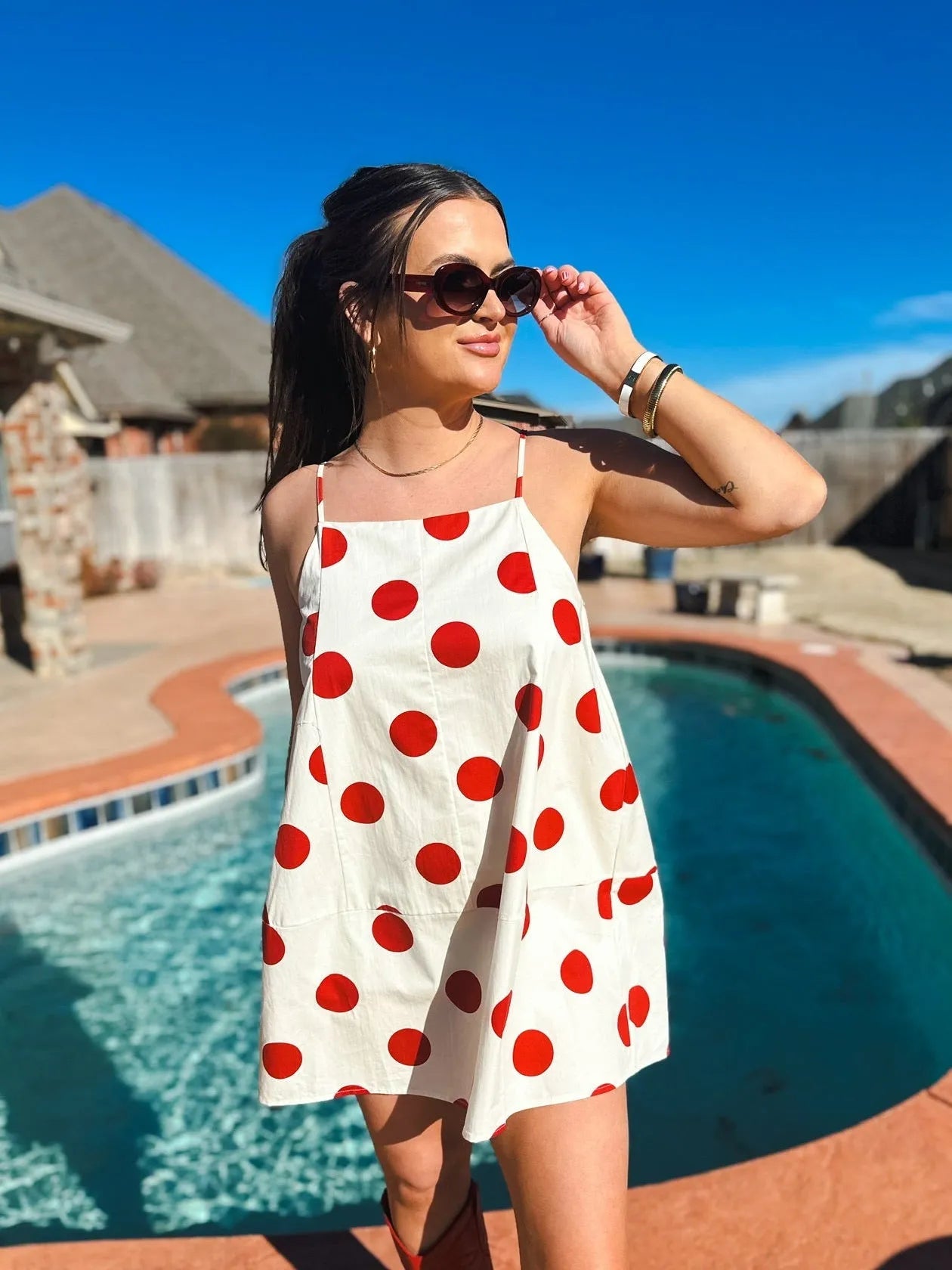 White mini dress with big red polka dots and spaghetti straps. Woman wearing sunglasses and gold necklace.