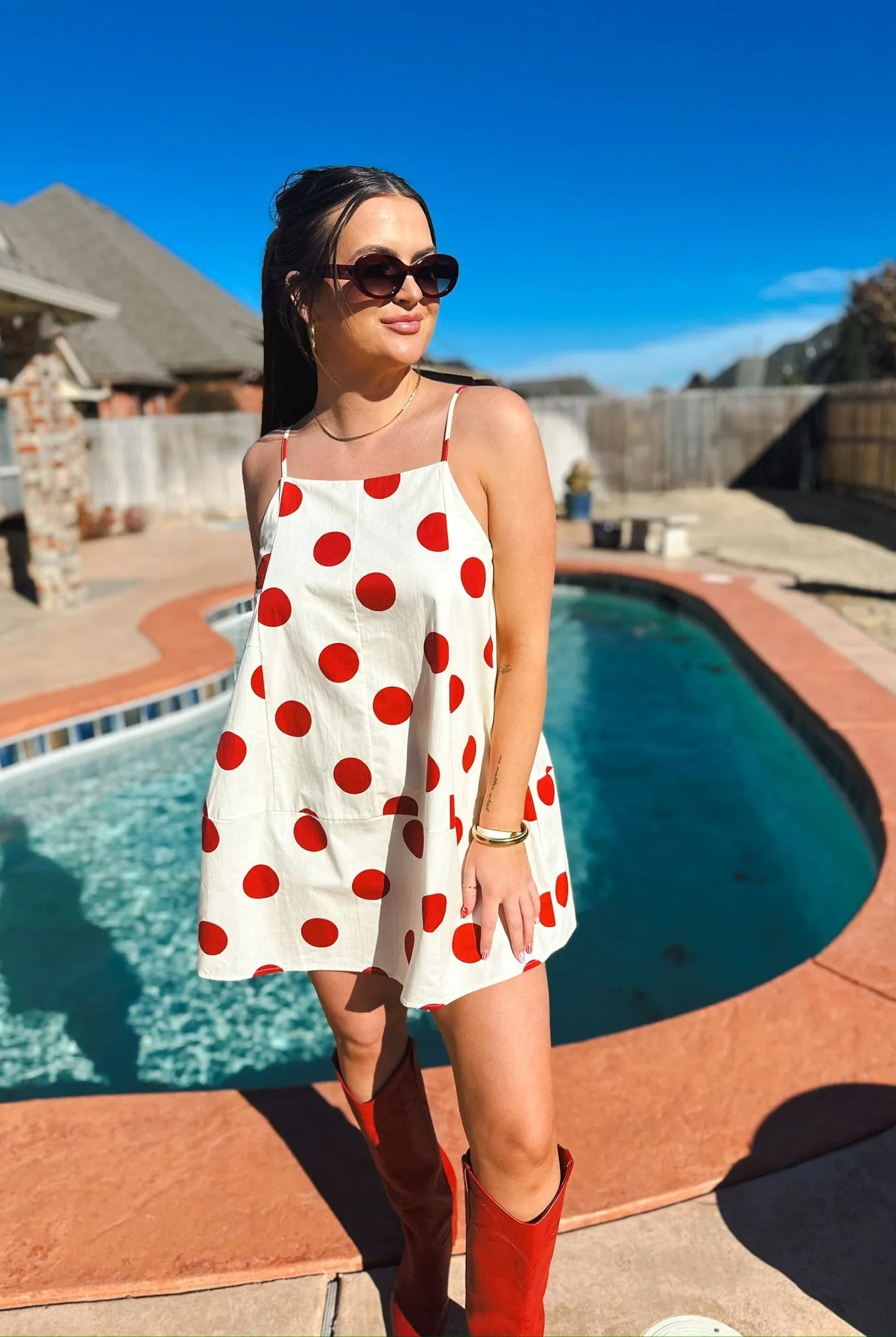 Woman in a white mini dress with big red polka dots and spaghetti straps. Red boots and sunglasses complete the look.