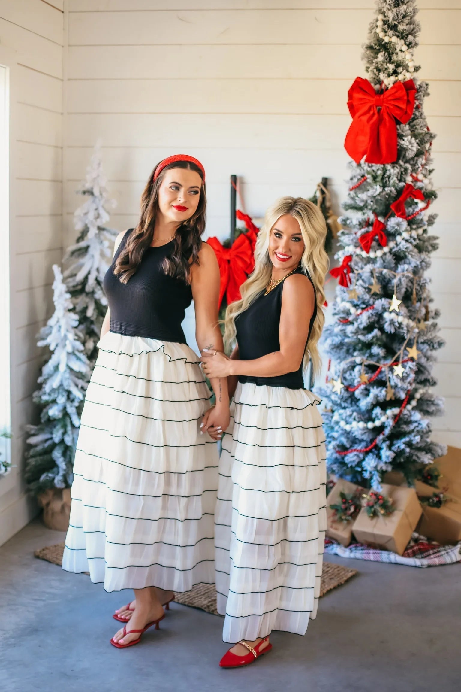 Two women in black tops and long, ivory ruffle dresses with black trim, posing near a decorated Christmas tree.