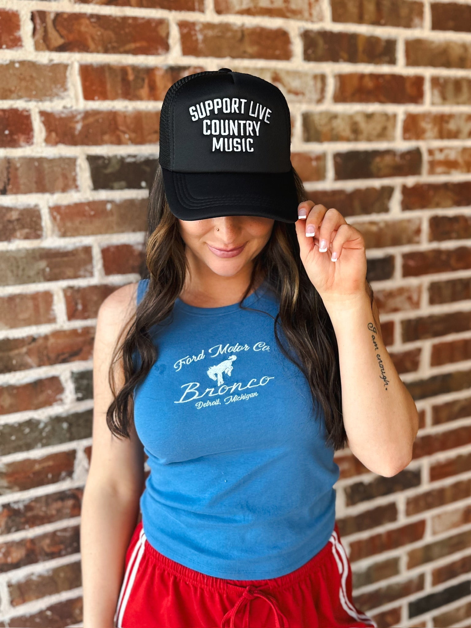 Black trucker hat with white text "SUPPORT LIVE COUNTRY MUSIC" on a brick background.