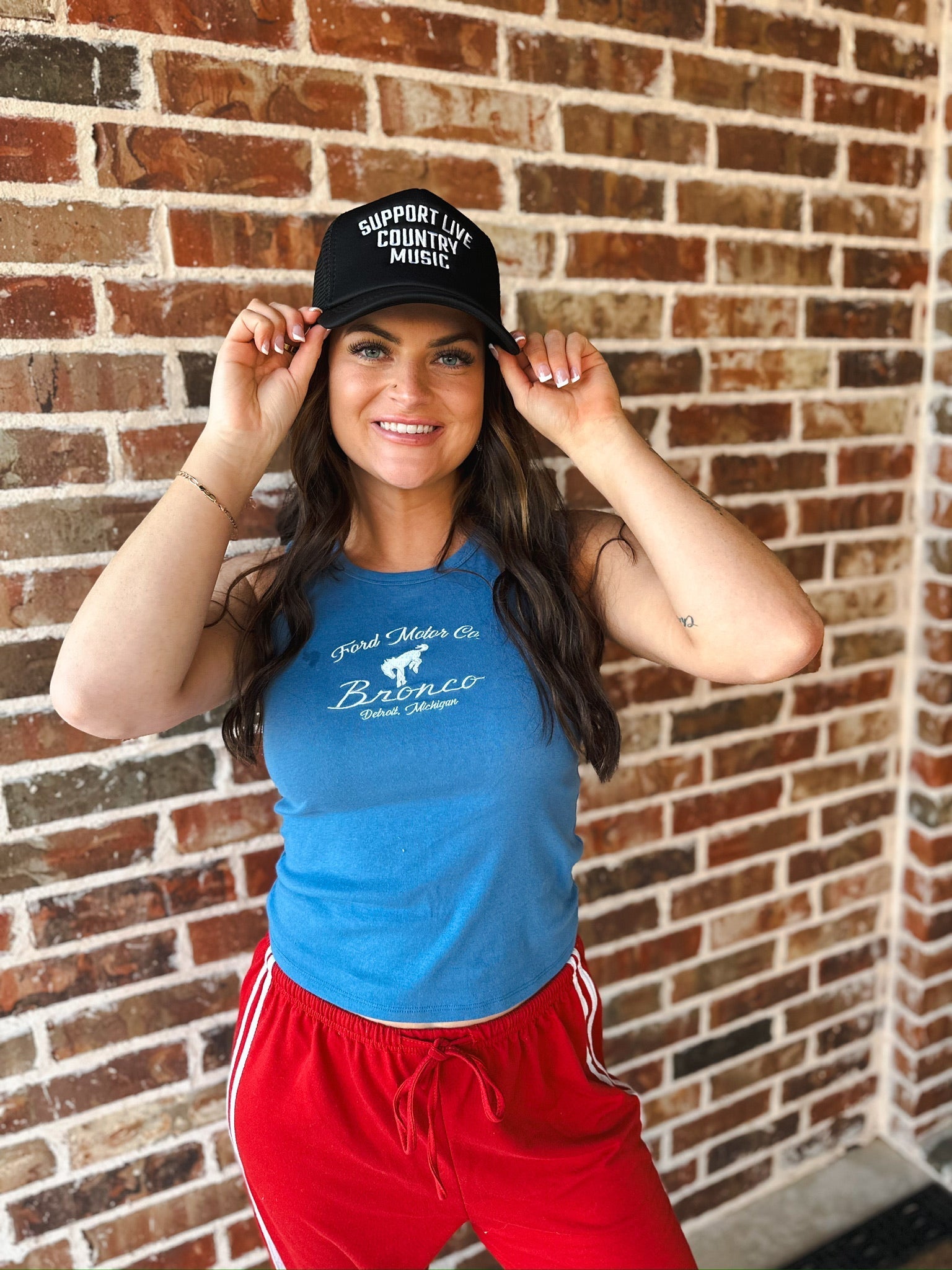 Black trucker hat with white text "SUPPORT LIVE COUNTRY MUSIC". Woman wearing blue Broncos tank top and red sweatpants.