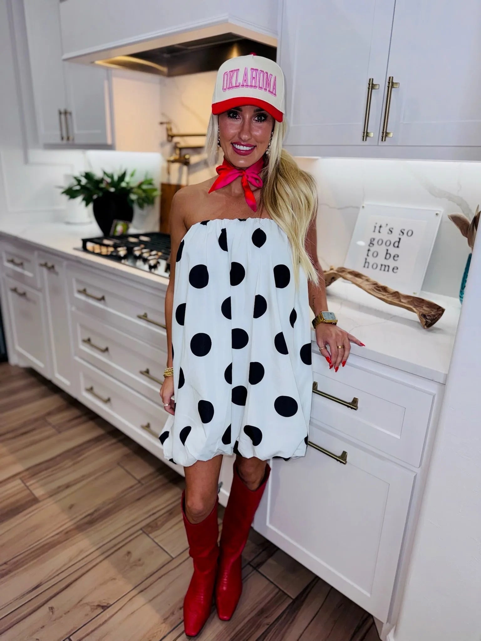 Woman in a strapless white dress with black polka dots, red cowboy boots, and an "Oklahoma" baseball cap.