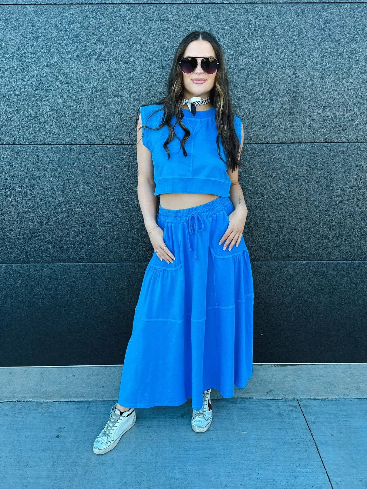 Woman wearing a vibrant blue sleeveless crop top and a tiered, flowing maxi skirt. Includes sunglasses and white sneakers.