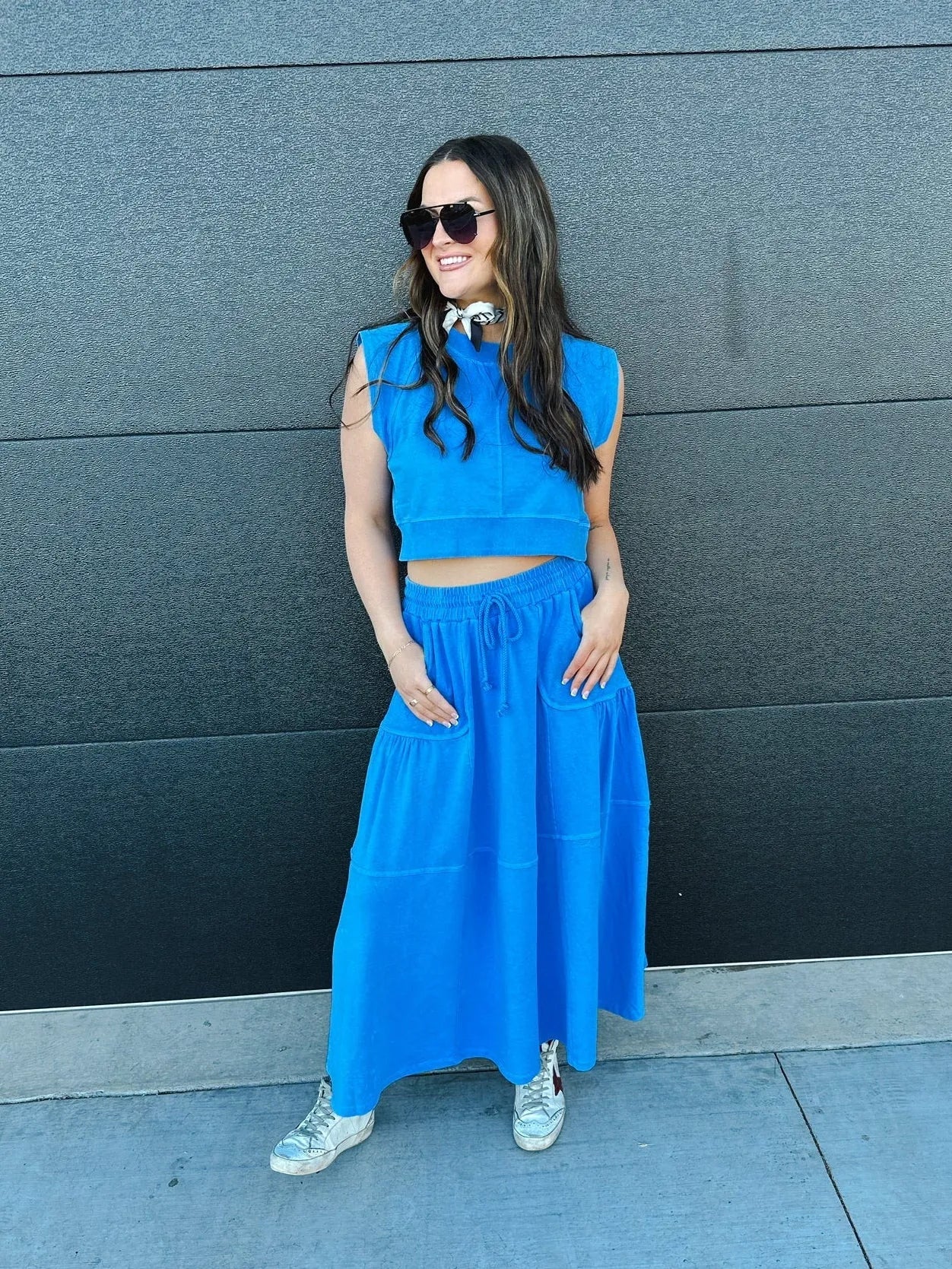 Woman wearing a vibrant blue sleeveless crop top and a tiered maxi skirt with pockets. She accessorizes with sunglasses and
