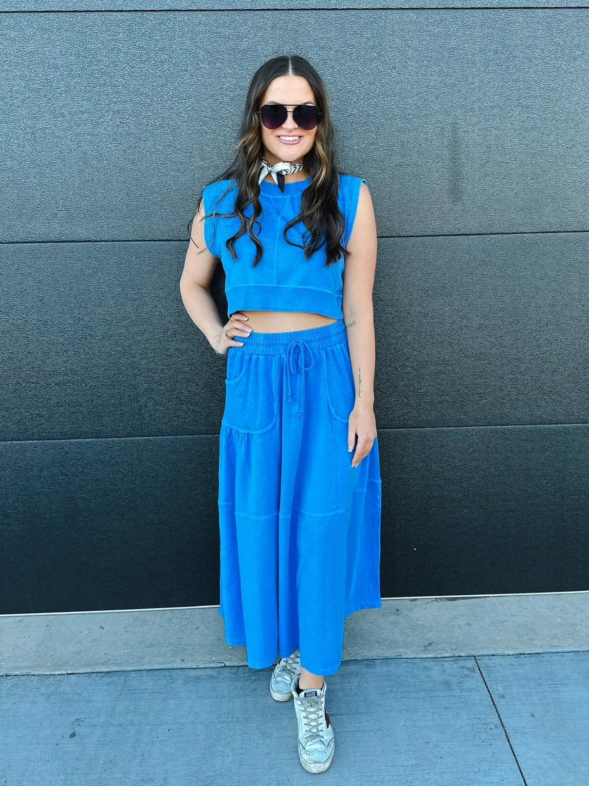 Woman in bright blue sleeveless crop top and tiered maxi skirt with drawstring waist, paired with sneakers and sunglasses.