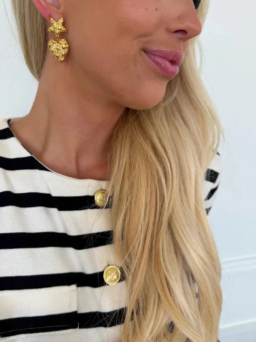 Gold star and textured heart drop earrings dangle from a model wearing a black and white striped shirt.