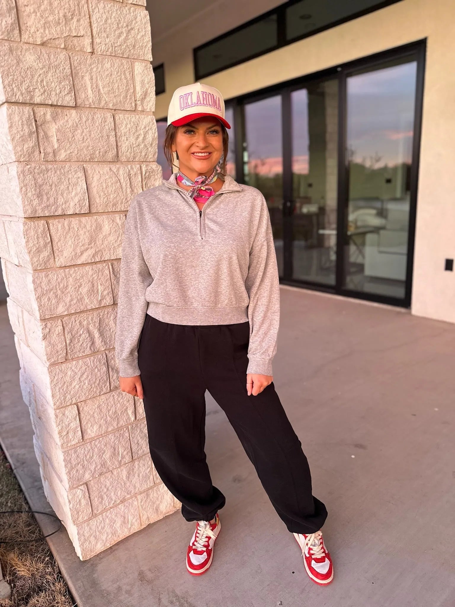 Model wears a grey collared half-zip sweatshirt, black joggers, and red and white sneakers. She is also wearing an