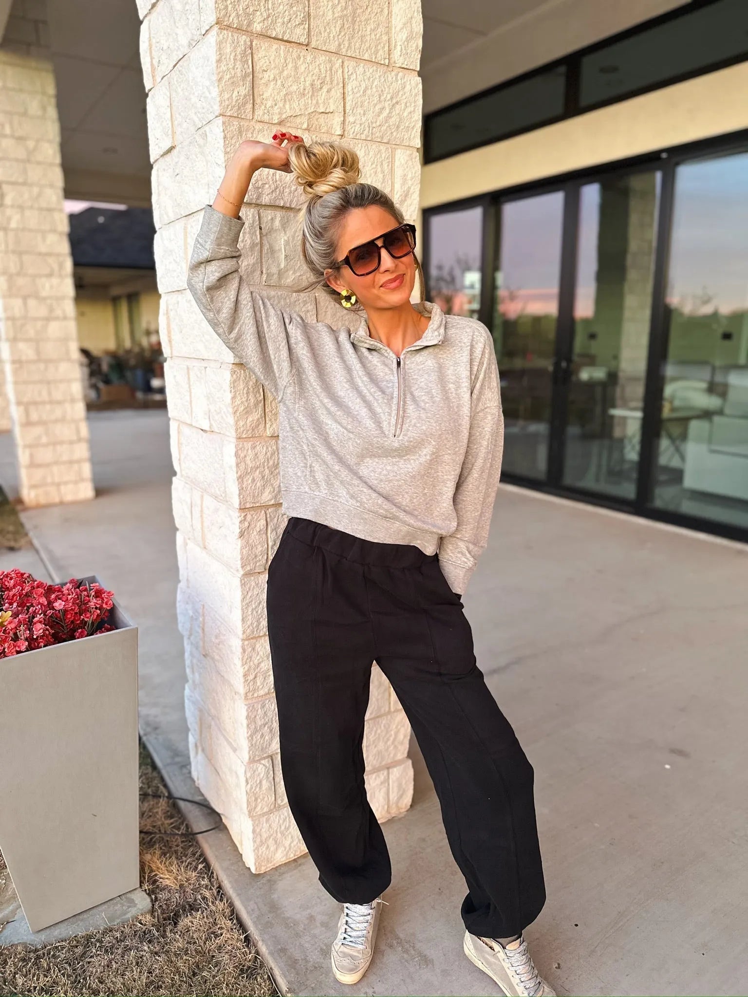 Woman wearing a light gray collared half-zip sweatshirt and black casual pants, leaning against a textured stone pillar