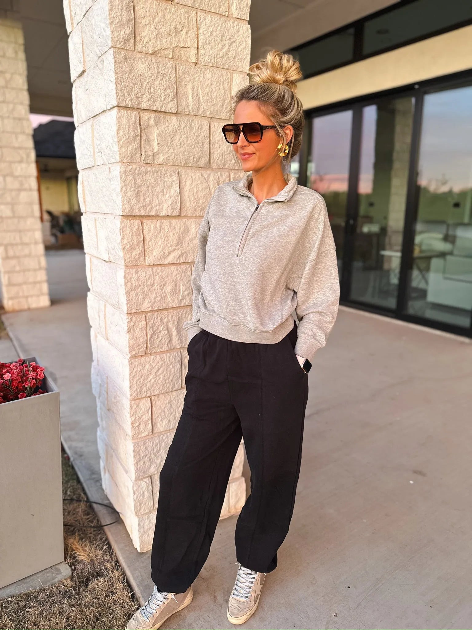 Woman wearing a grey collared half-zip sweatshirt and black pants, leaning against a brick pillar.
