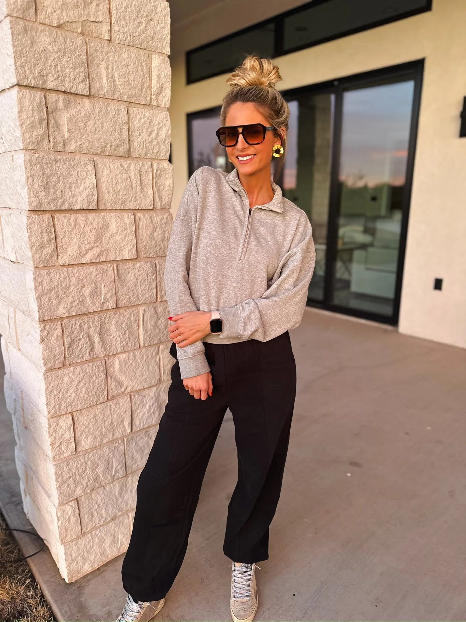 Woman wearing a grey collared half-zip sweatshirt, black pants, and sneakers. She has blonde hair in a bun, sunglasses, and