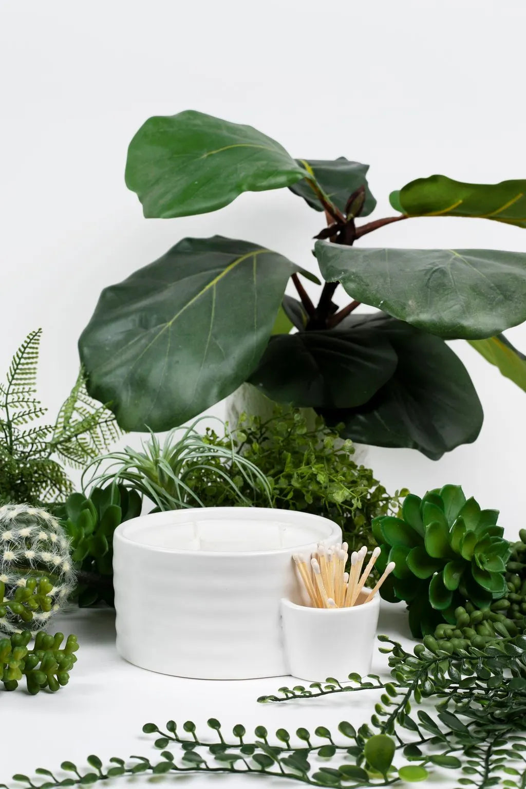 White ceramic Palm Desert match holder candle with three wicks, surrounded by various green plants and a pot of matches.