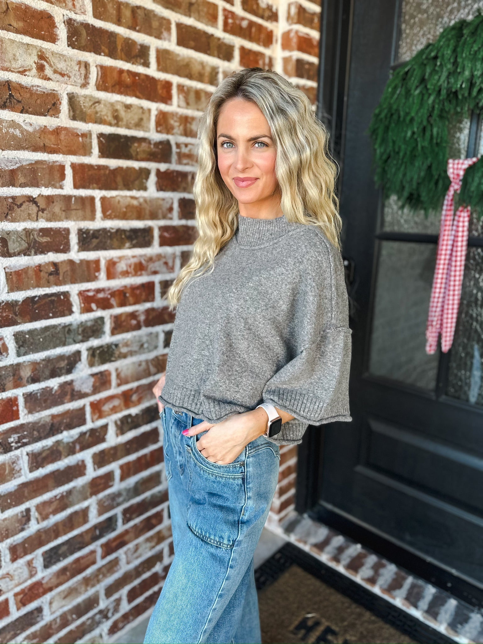Woman wearing a charcoal brown soft knit sweater tee with wide sleeves and blue jeans, standing by a brick wall.