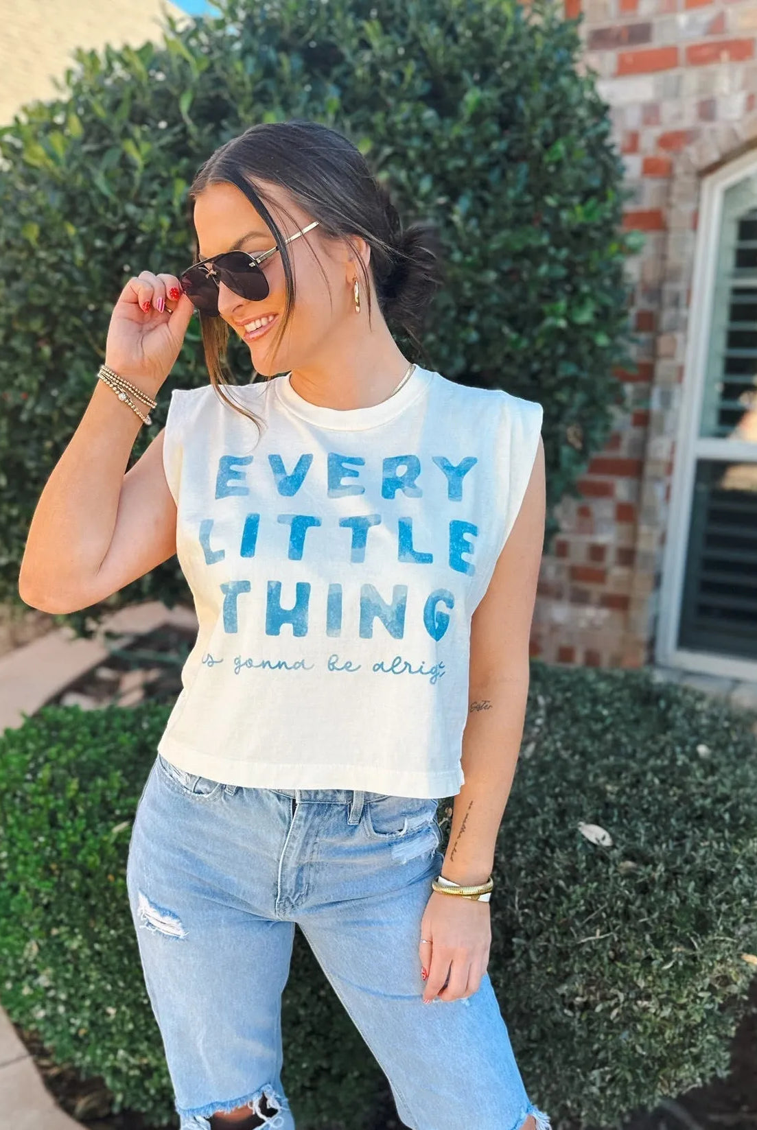 Woman wearing a white casual tank top with blue text that says 'EVERY LITTLE THING is gonna be alright' and light wash