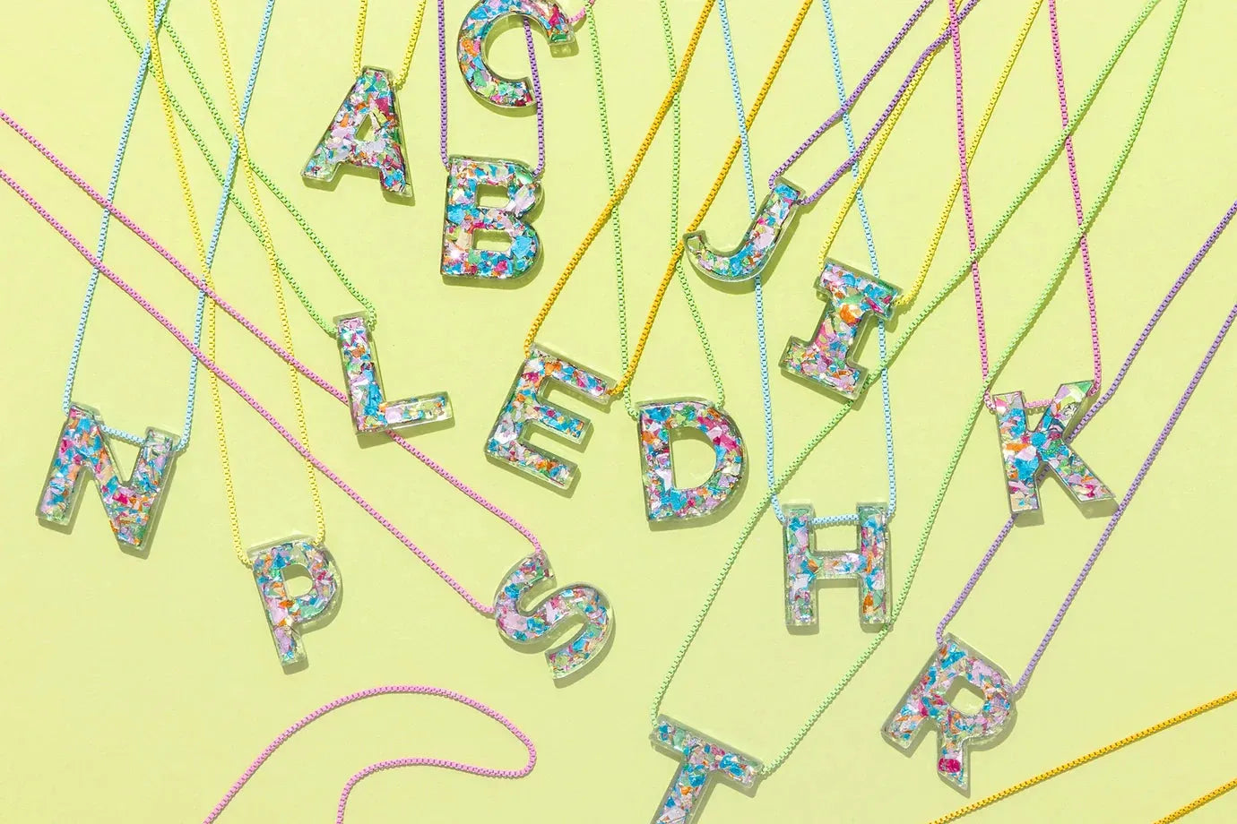 Confetti initial necklaces with letters A, B, C, D, E, F, H, I, J, K, L, N, O, P, R, S, T displayed on a light yellow