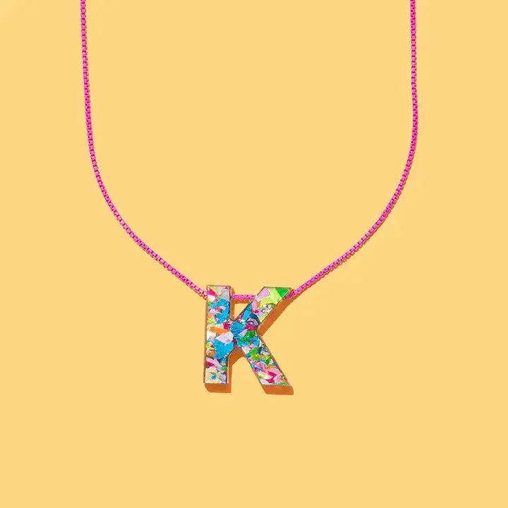 Confetti initial K necklace with a bright pink chain on a yellow background. The letter K is filled with colorful, abstract