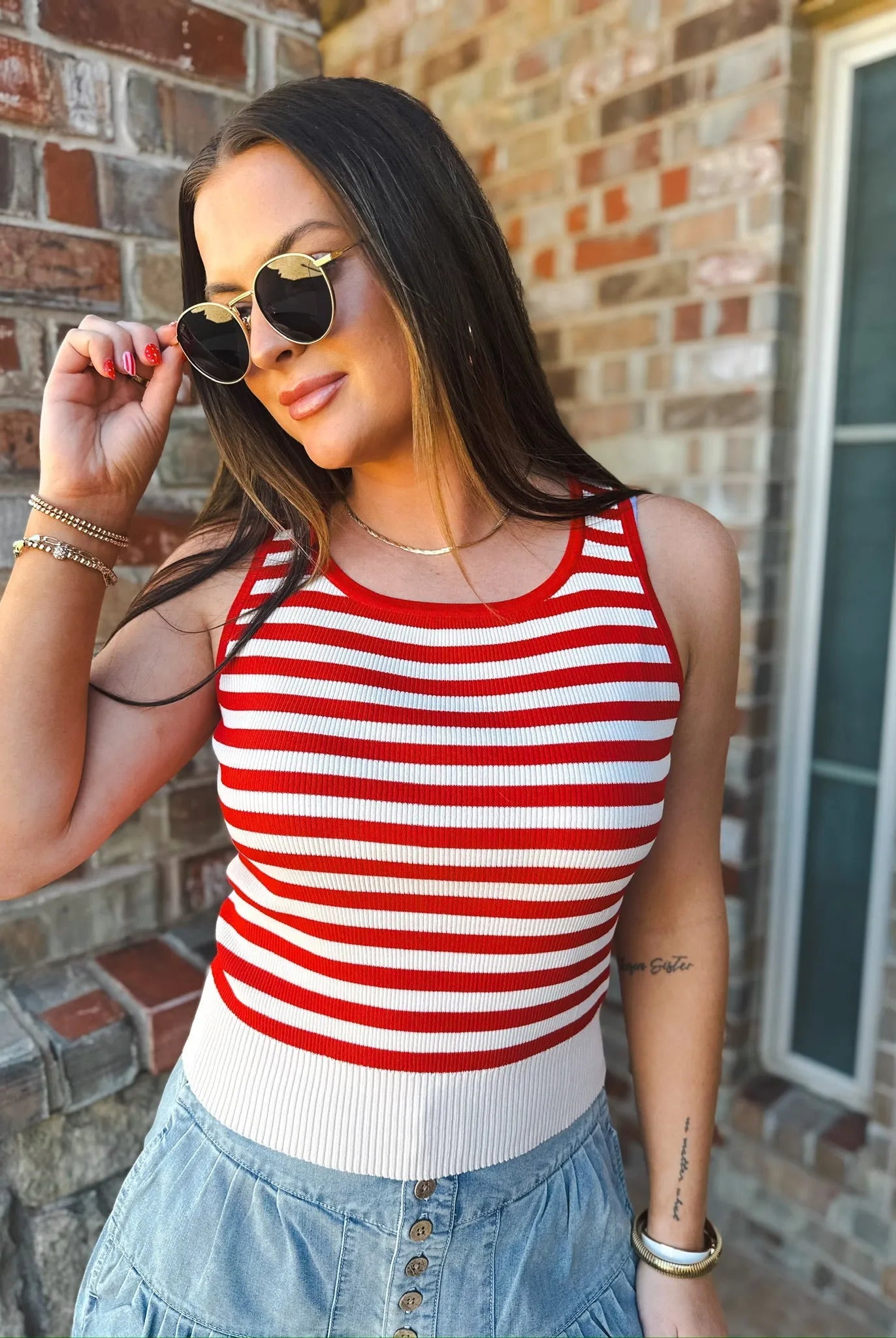 Woman wearing a cream and red stripe game day tank top and denim skirt, adjusting sunglasses.
