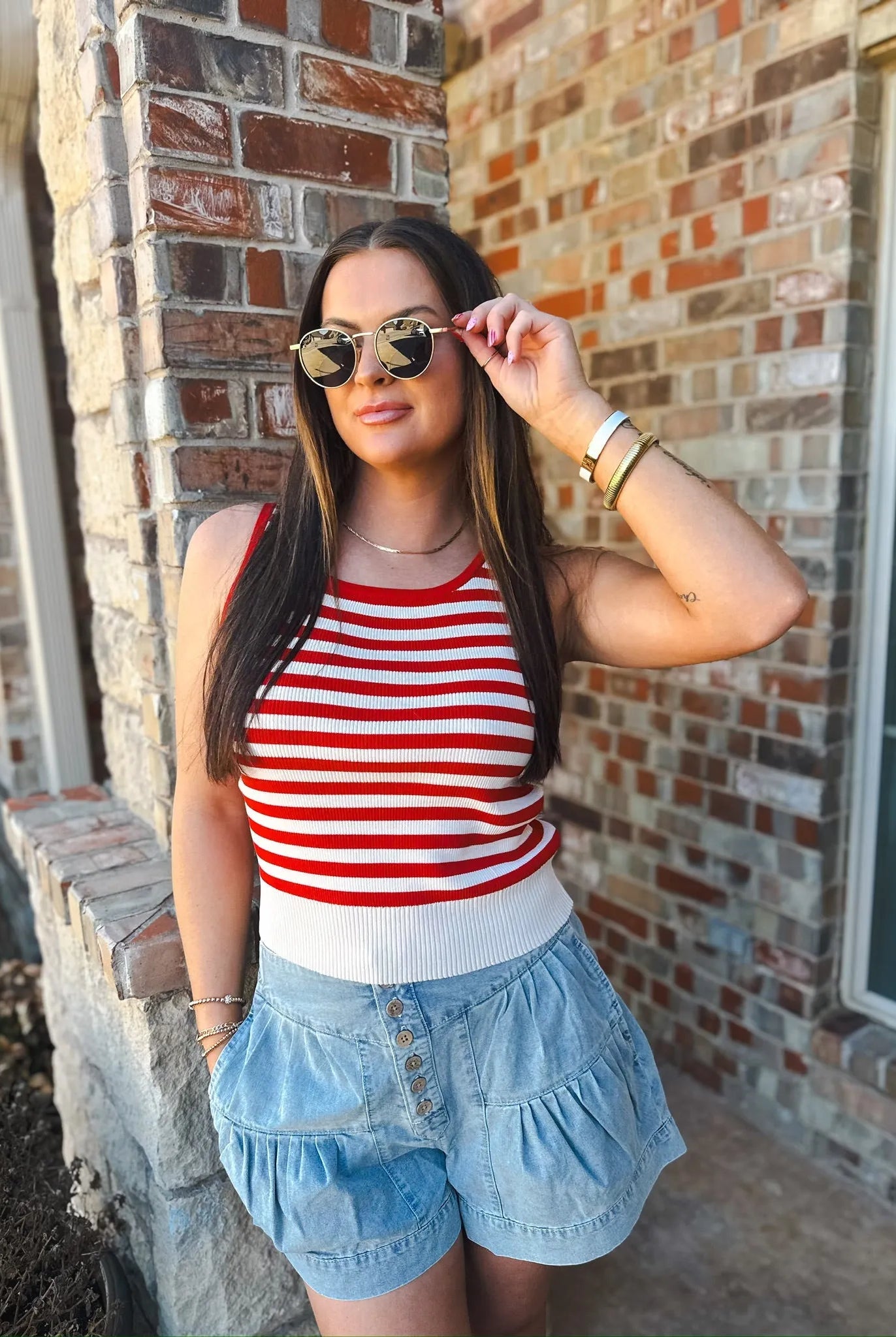 Woman wearing a red and cream striped knit tank top and light wash denim ruffle shorts, accessorized with sunglasses and